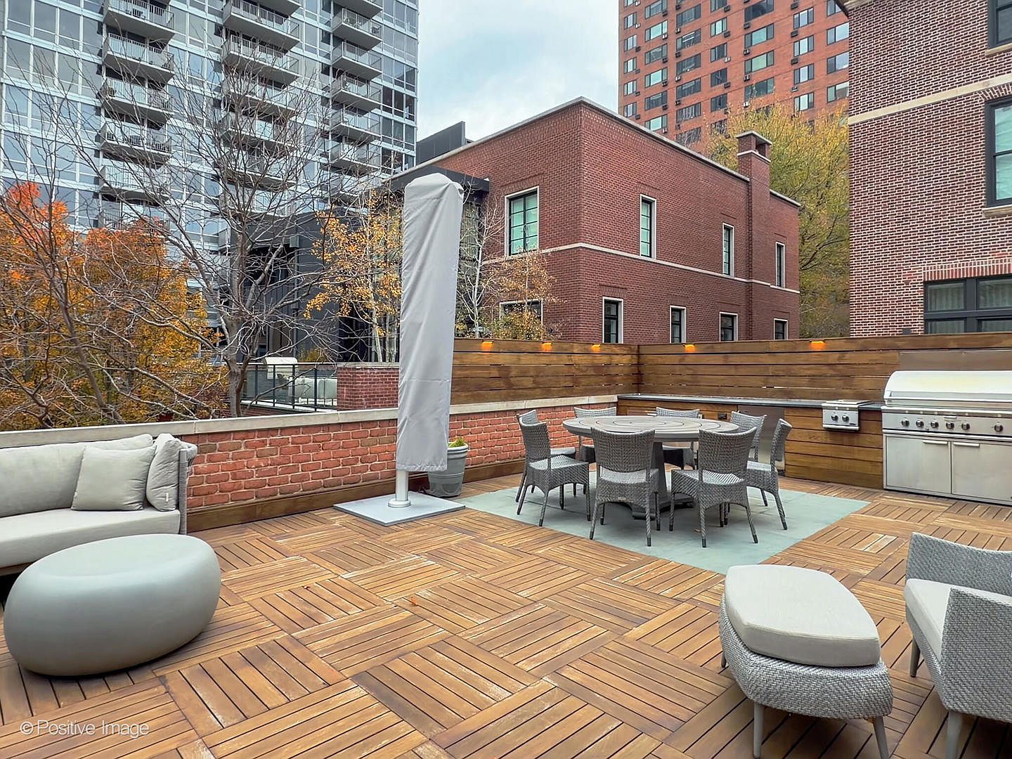This expansive rooftop terrace features high-end wood decking arranged in a modern parquet pattern, creating a sophisticated outdoor living space. The area is well-appointed with a comfortable lounge seating group, a dining set for six, and a built-in stainless steel grill station, all framed by a contemporary horizontal wood fence. The perspective captures a serene urban oasis, blending luxury amenities with a backdrop of city architecture and autumn foliage.