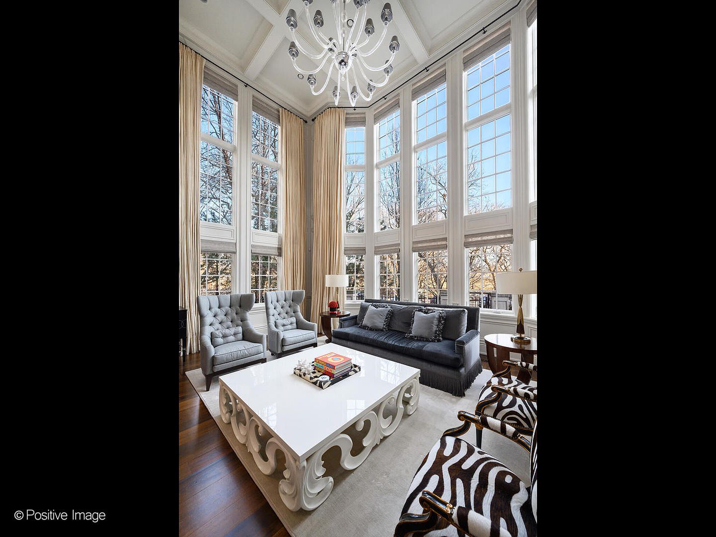 This grand, double-height living room exudes luxury with its soaring ceilings, floor-to-ceiling windows, and an ornate, multi-armed chandelier. The space is anchored by a large, white, intricately carved coffee table, surrounded by a plush grey sofa, two tufted wingback chairs, and a pair of stylish zebra-print accent chairs. The room's sophisticated design, featuring rich hardwood floors and elegant drapery, creates an airy and opulent atmosphere perfect for formal entertaining.