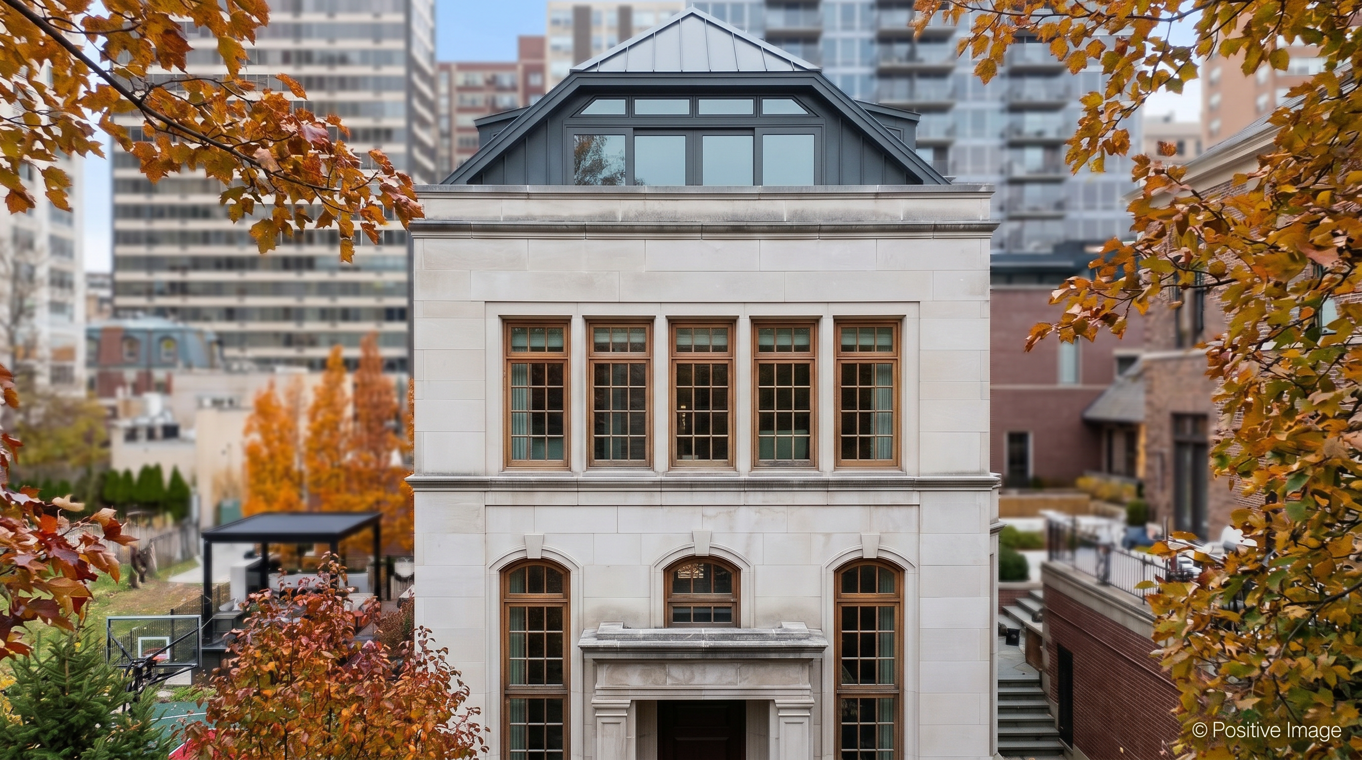 This elegant limestone residence features a symmetrical neoclassical design characterized by tall, wood-framed windows and a distinct gabled upper level with a dark metal roof. The facade is meticulously detailed with stone trim, while the surrounding property is framed by vibrant autumn foliage, offering a sophisticated and tranquil suburban impression.