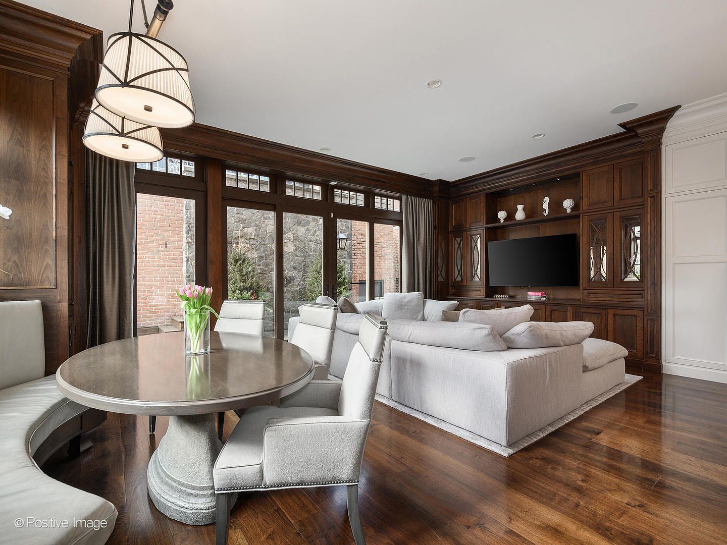 This elegant open-concept living and dining space features rich, dark wood paneling and cabinetry that contrast beautifully with the light-toned, plush sectional sofa and dining chairs. A circular dining table with a stone pedestal base sits near a large window wall that provides a view of a stone-walled courtyard, while a large television is integrated into the custom built-in shelving. The room exudes a sophisticated, warm, and inviting atmosphere, perfect for both relaxation and formal entertaining.