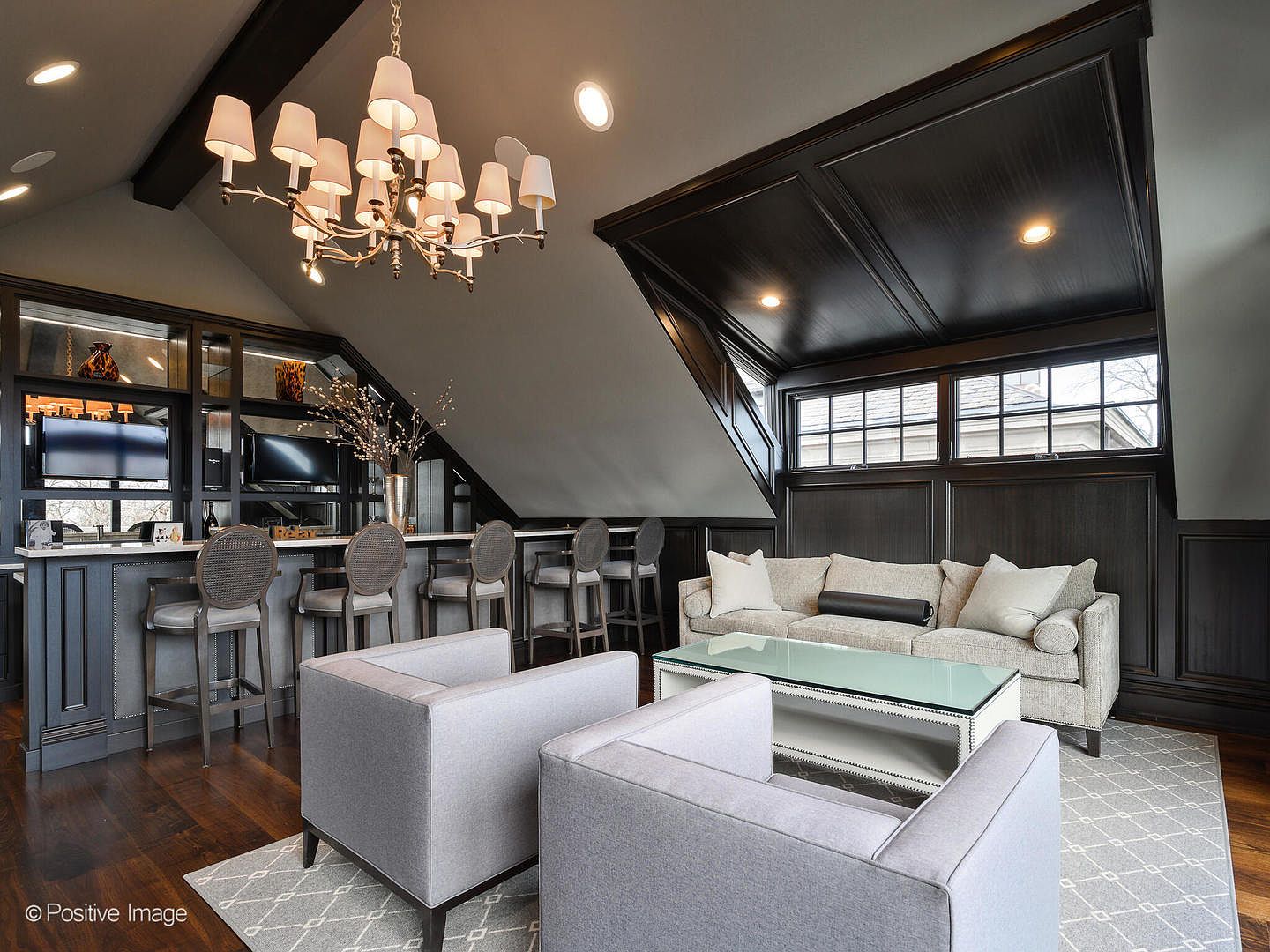 This sophisticated attic lounge features a built-in bar with dark cabinetry, a cozy seating area with a light-toned sofa and two armchairs, and a glass-topped coffee table. The space is defined by its dramatic sloped ceiling with dark wood paneling, a classic chandelier, and large windows that bring in natural light. The overall aesthetic is elegant and refined, creating a perfect retreat for relaxation or entertaining.