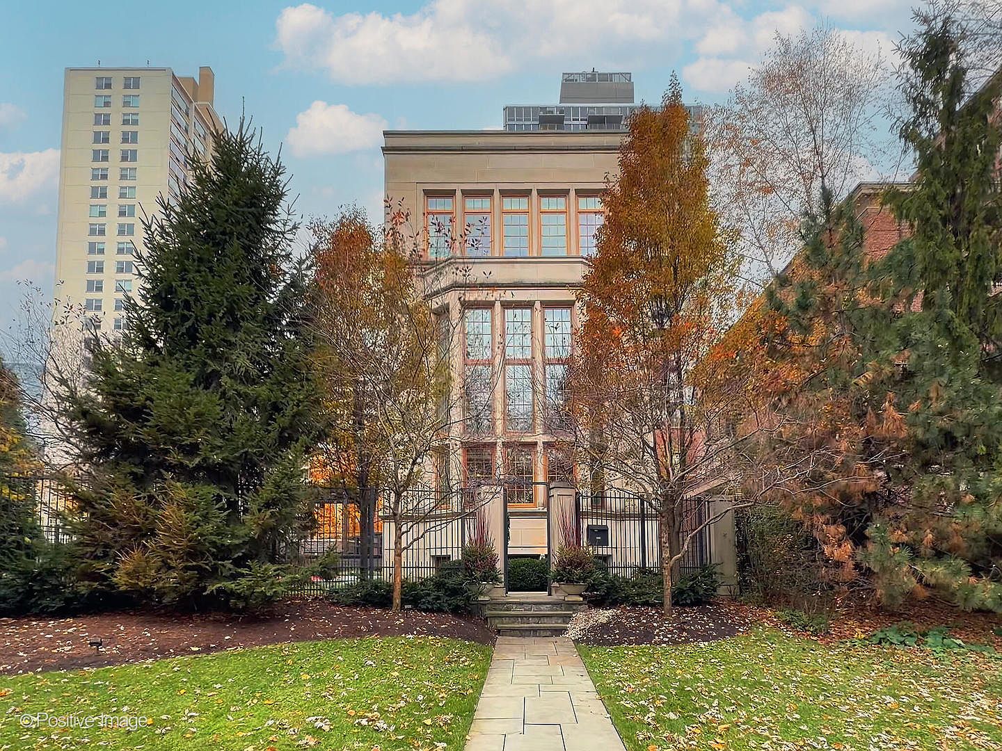 This elegant, multi-story stone residence features a symmetrical facade with large, grid-paned windows that create a sophisticated, classic aesthetic. A stone pathway leads through a manicured lawn and mature trees toward a gated entrance, framed by wrought-iron fencing. The perspective is a straight-on, eye-level shot that emphasizes the grand scale and architectural symmetry of the home against a backdrop of a clear sky and a neighboring high-rise.