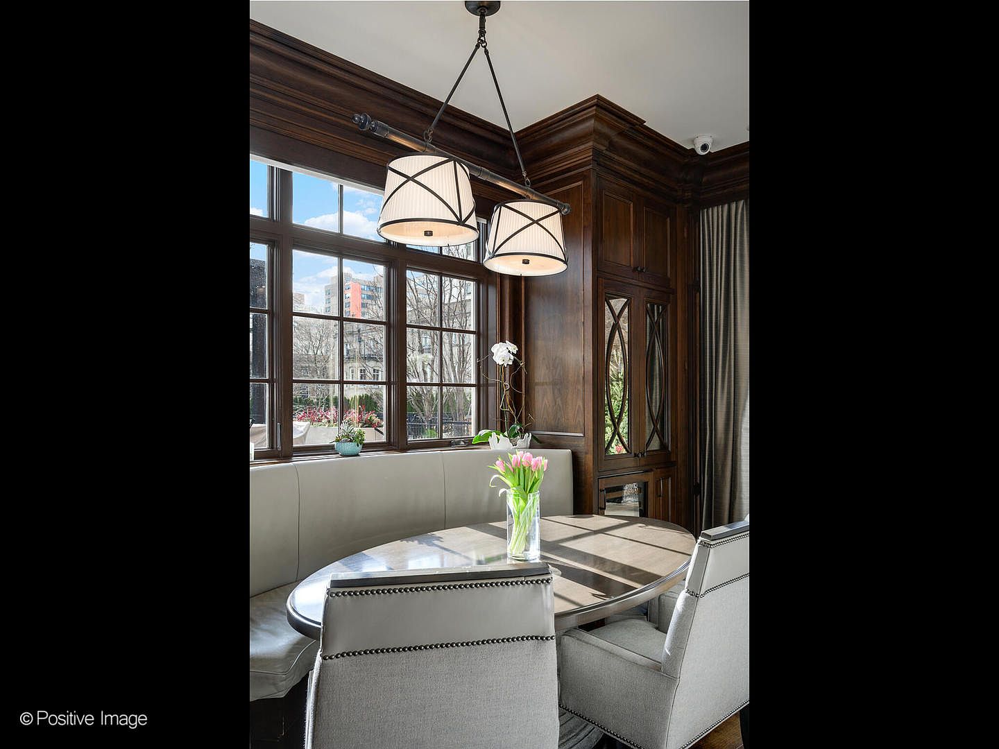 This elegant dining nook features a built-in banquette upholstered in light gray fabric, paired with a round table and matching chairs. The space is framed by rich, dark wood paneling and large windows that allow natural light to illuminate the room, while a stylish dual-pendant light fixture hangs above. The overall aesthetic is sophisticated and cozy, perfect for intimate meals in a high-end residential setting.