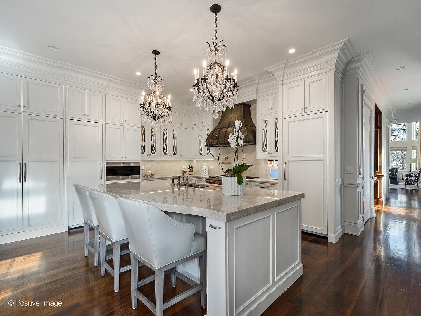This elegant, high-end kitchen features crisp white cabinetry, a large central island with a stone countertop, and three upholstered bar stools. The space is illuminated by two ornate crystal chandeliers, while a dark, custom-built range hood serves as a dramatic focal point above the stove. The warm hardwood flooring and classic architectural details create a sophisticated and inviting atmosphere, captured from a wide-angle perspective that emphasizes the room's spacious layout.