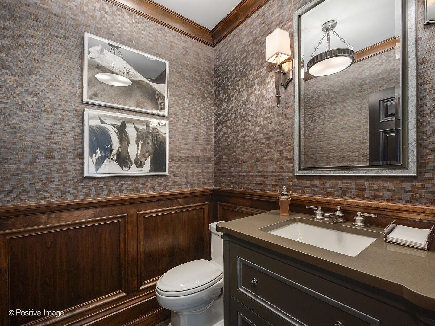 This sophisticated powder room features rich wood wainscoting paired with textured, metallic-toned wallpaper and two framed horse photographs. The space includes a dark vanity with a stone countertop, a modern undermount sink, and a sleek, framed mirror illuminated by a stylish wall sconce. The overall aesthetic is refined and masculine, creating a polished and inviting atmosphere for guests.