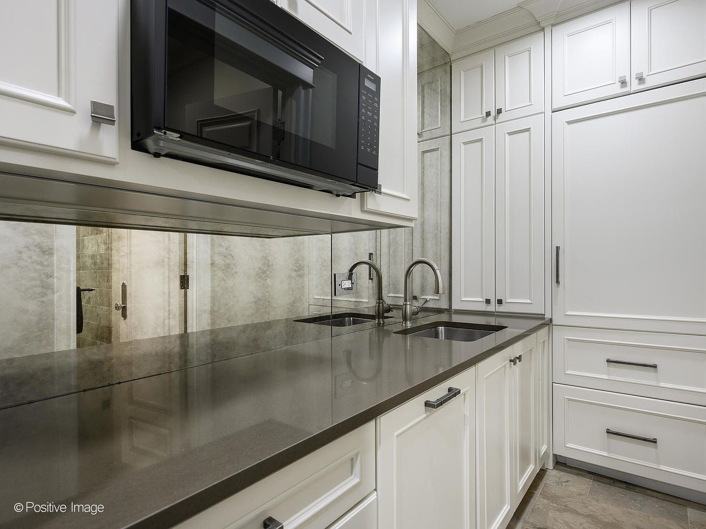 This sleek, modern kitchen area features crisp white cabinetry paired with a dark, polished stone countertop and a reflective mirrored backsplash that adds depth to the space. A built-in black microwave is mounted above the counter, which includes a stainless steel sink and a high-end faucet. The composition is captured from a straight-on, eye-level perspective, highlighting the clean lines and sophisticated, high-contrast aesthetic of the cabinetry and hardware.