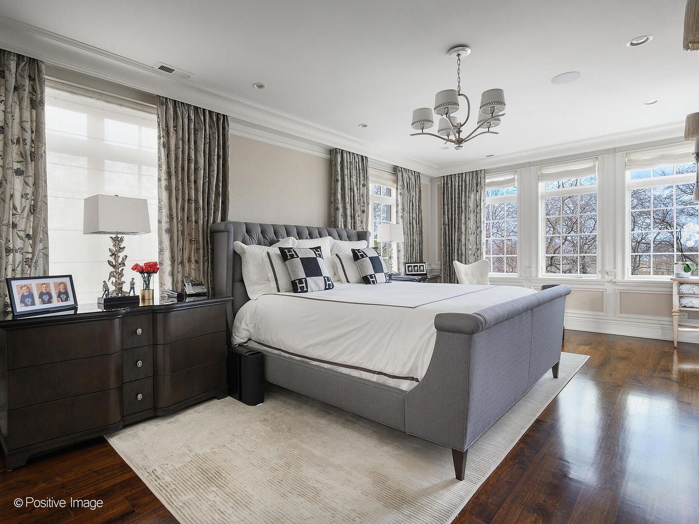 This elegant primary bedroom features a sophisticated gray tufted sleigh bed as the focal point, complemented by dark wood nightstands and a neutral color palette. Large windows draped in patterned curtains allow ample natural light to fill the room, highlighting the rich hardwood floors and the classic chandelier above. The space exudes a refined, tranquil atmosphere, perfect for a luxury residential listing.