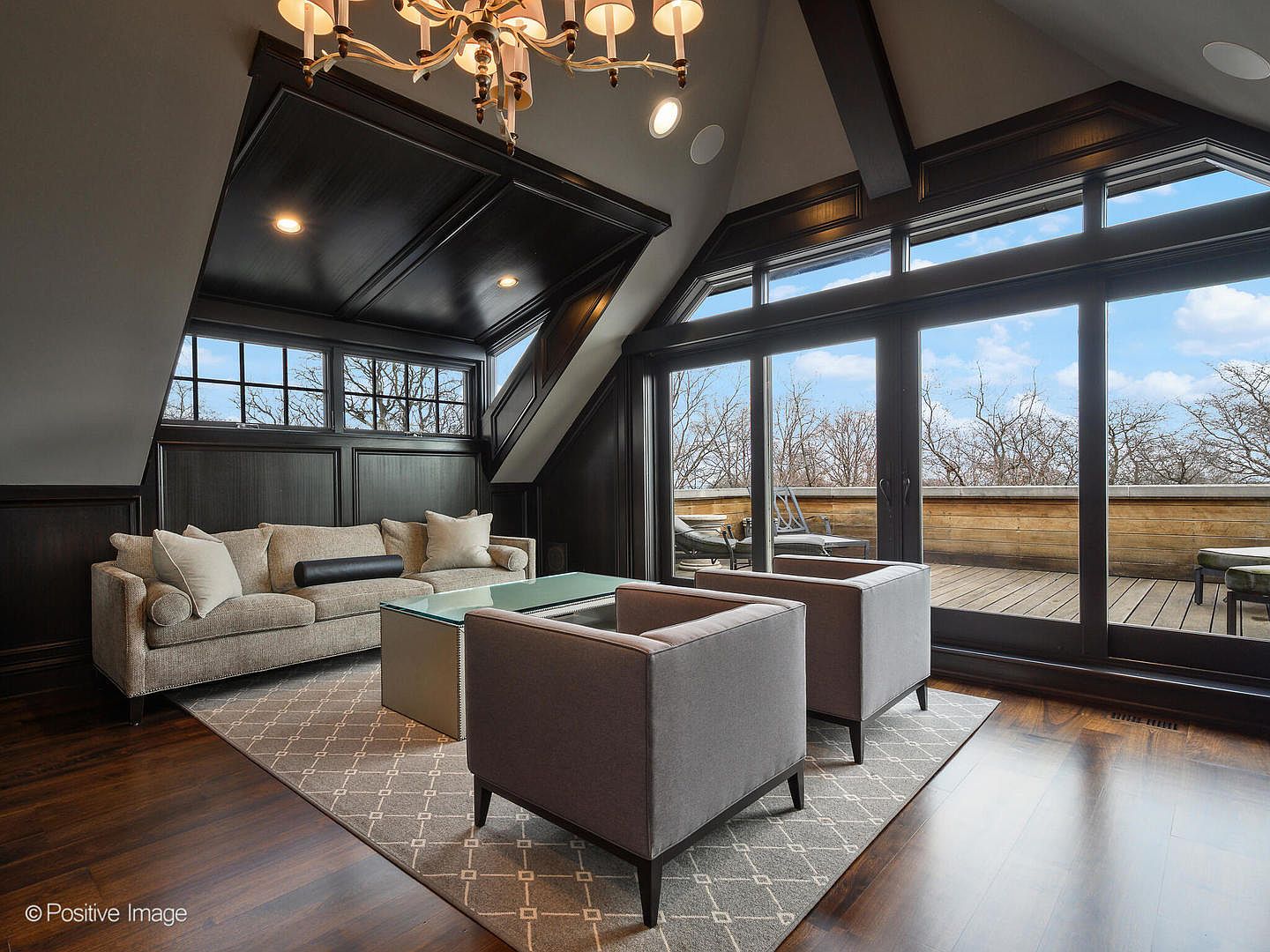 This sophisticated living room features a dramatic vaulted ceiling with dark wood paneling and a classic chandelier, creating a luxurious and cozy atmosphere. The space is furnished with a plush beige sofa and two modern grey armchairs arranged on a geometric patterned rug, all centered around a sleek glass-topped coffee table. Large floor-to-ceiling sliding glass doors provide an expansive view of an outdoor deck, seamlessly blending indoor comfort with the natural surroundings.