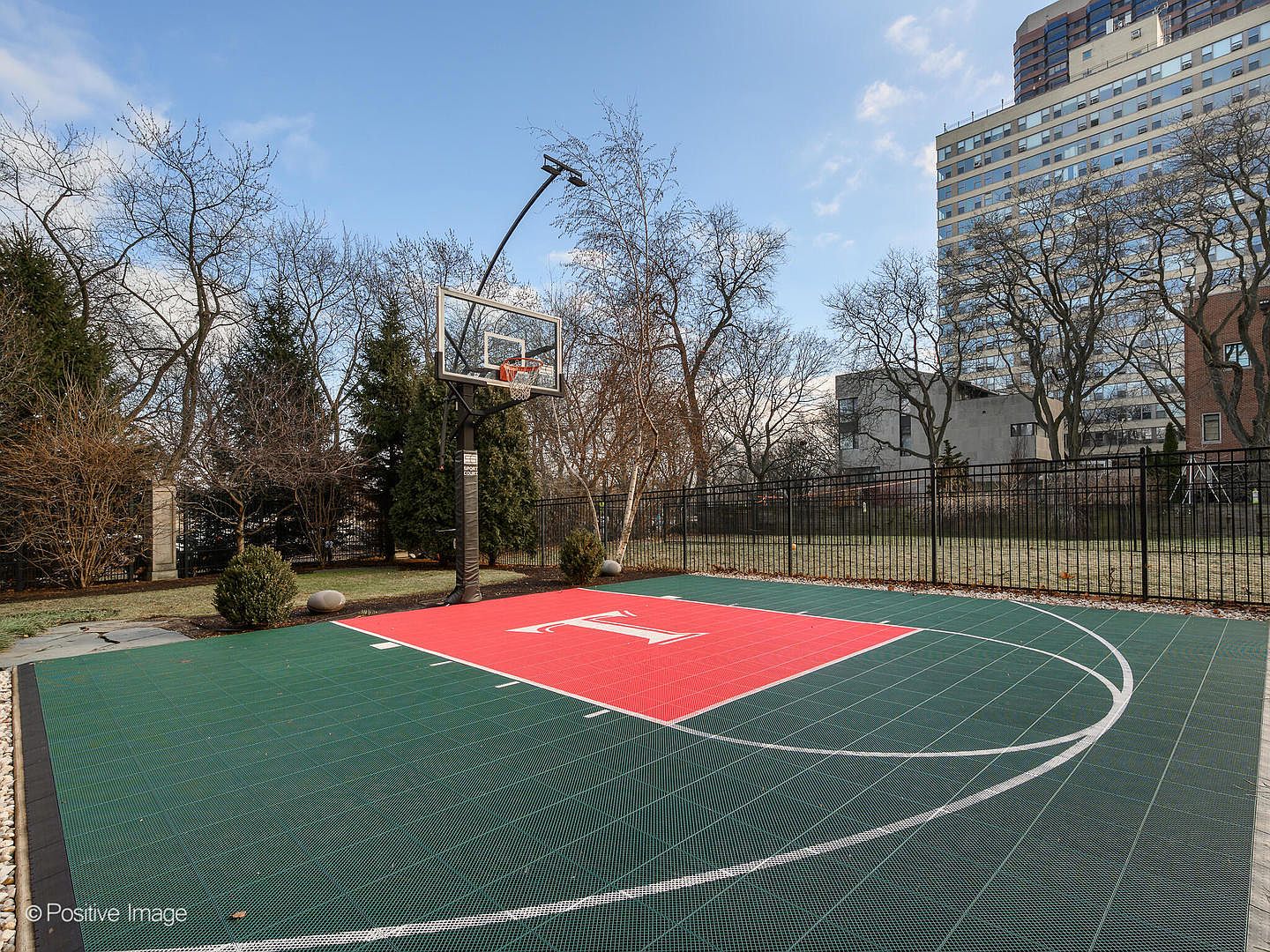 This image showcases a private, professional-grade basketball court featuring a vibrant green and red modular surface with a prominent white 'T' logo. The court is enclosed by a black metal fence and is framed by mature, leafless trees, with a large residential high-rise building visible in the background. The perspective is a wide-angle shot from the court level, emphasizing the recreational space's integration into the surrounding landscape.
