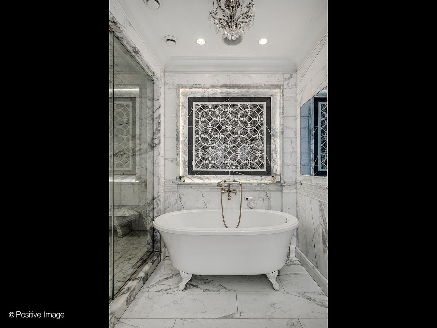 This luxurious primary bathroom features a classic white clawfoot tub centered against a striking marble wall with an intricate geometric mosaic inlay. The space is adorned with elegant marble tiling throughout, a crystal chandelier, and a glass-enclosed shower, creating a sophisticated and spa-like atmosphere. The symmetrical composition and high-end finishes highlight the room's opulent design.