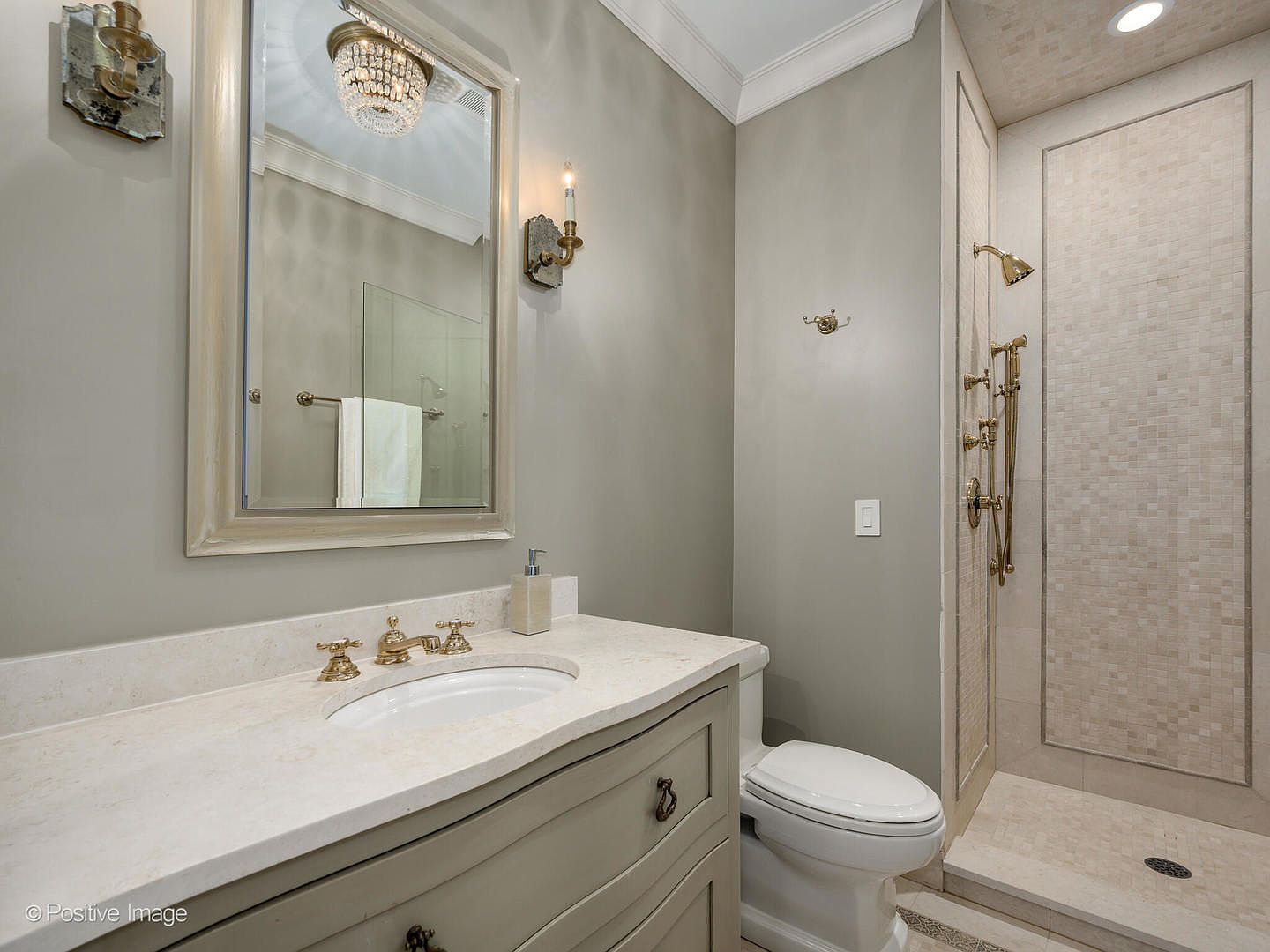 This elegant bathroom features a light gray vanity with a stone countertop, a framed mirror with a crystal chandelier reflection, and a walk-in shower with mosaic tile accents. The space is finished with polished gold fixtures and neutral wall tones, creating a sophisticated and clean aesthetic. The perspective captures the vanity and shower area, highlighting the high-end finishes and compact, functional layout.