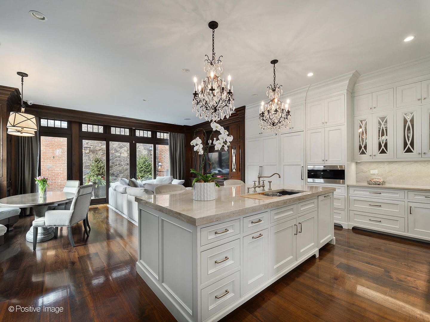 This elegant, high-end kitchen features a large central island with a stone countertop, white cabinetry, and two ornate crystal chandeliers that create a sophisticated focal point. The space seamlessly transitions into an adjacent dining area and living room, framed by dark wood-trimmed windows that overlook a stone-walled patio. The combination of classic architectural details, rich hardwood floors, and bright, clean finishes provides a luxurious and inviting atmosphere.