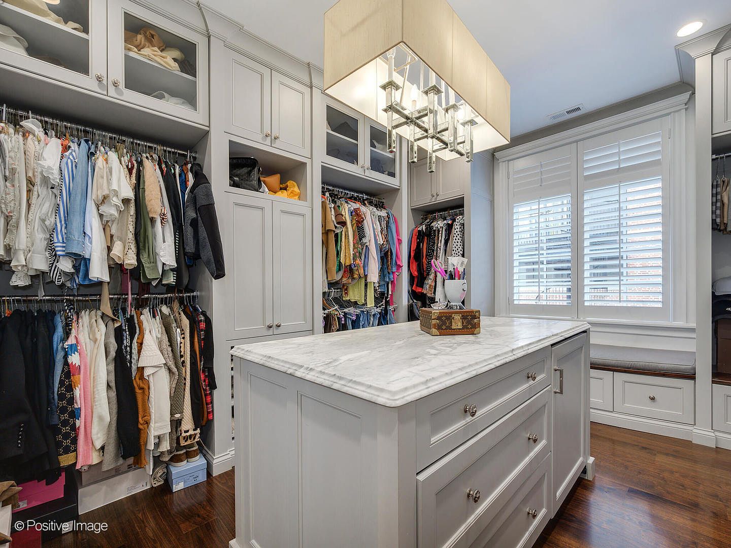 This luxurious walk-in closet features custom light-gray cabinetry with ample hanging space and glass-fronted upper storage. A central island with a white marble countertop serves as a sophisticated focal point, complemented by a modern rectangular chandelier overhead. The space is finished with rich hardwood flooring and a built-in window seat, creating an elegant and organized dressing area.