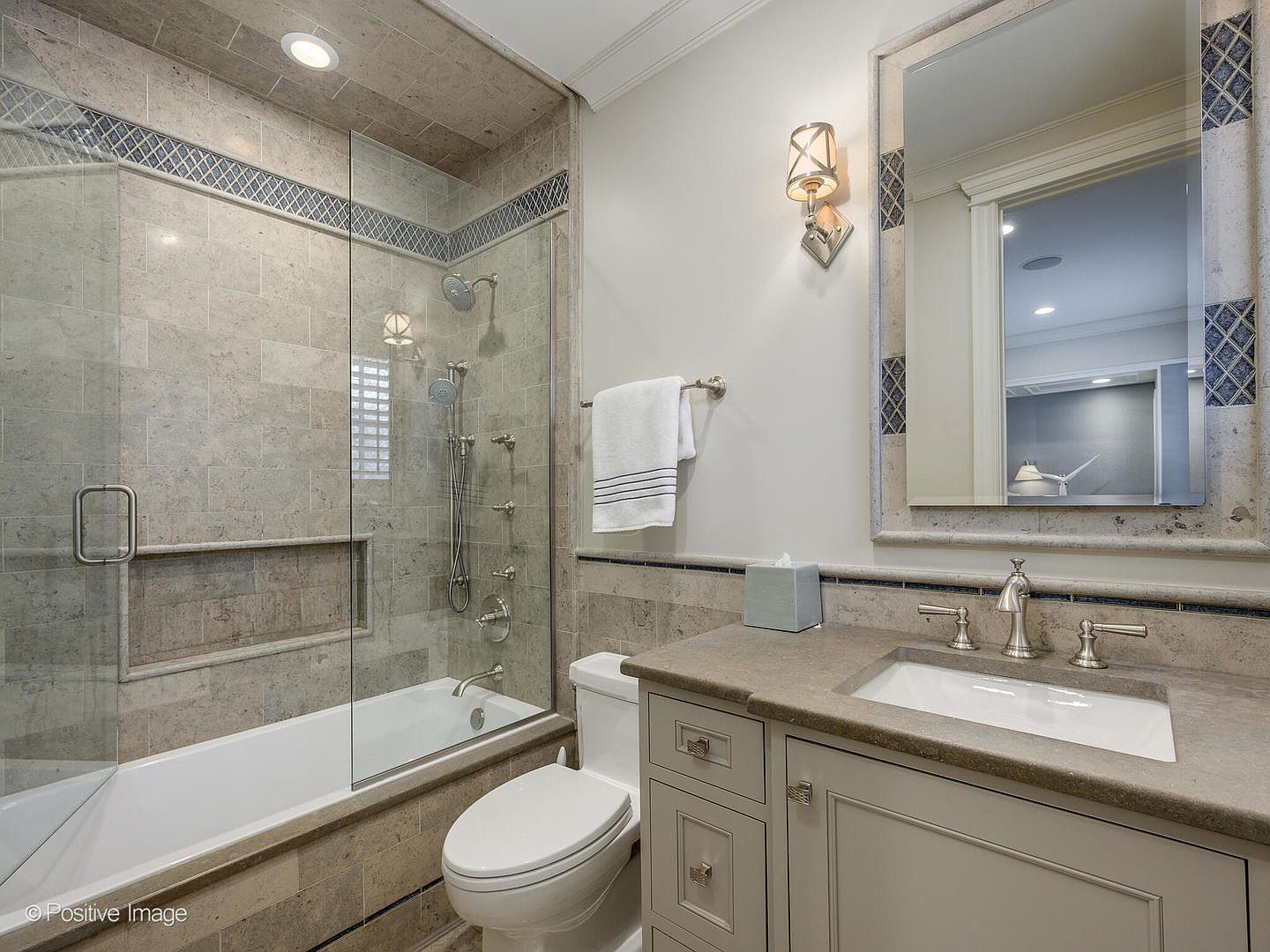 This well-appointed bathroom features a neutral color palette with stone-tiled walls and a glass-enclosed shower-tub combination. The vanity includes a stone countertop with an undermount sink, complemented by a framed mirror and elegant wall sconce lighting. The space is clean, functional, and designed with a classic, sophisticated aesthetic suitable for a guest or secondary bathroom.