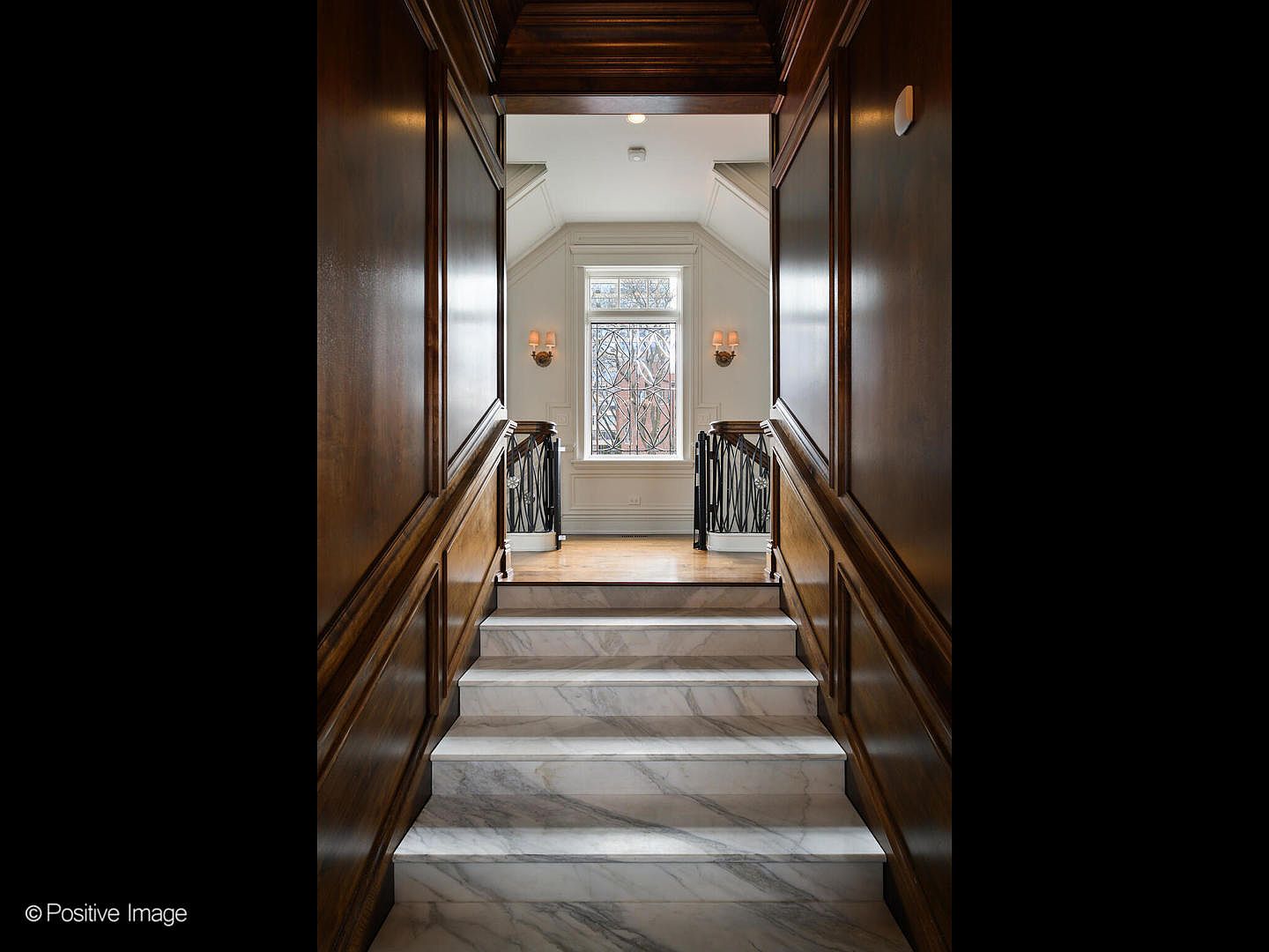 This elegant staircase features luxurious white marble steps flanked by rich, dark wood-paneled walls that create a sophisticated and dramatic entryway. The perspective looks upward toward a landing illuminated by a large, decorative window with intricate ironwork, flanked by classic wall sconces. The combination of warm wood tones and cool stone creates a high-end, timeless architectural aesthetic.