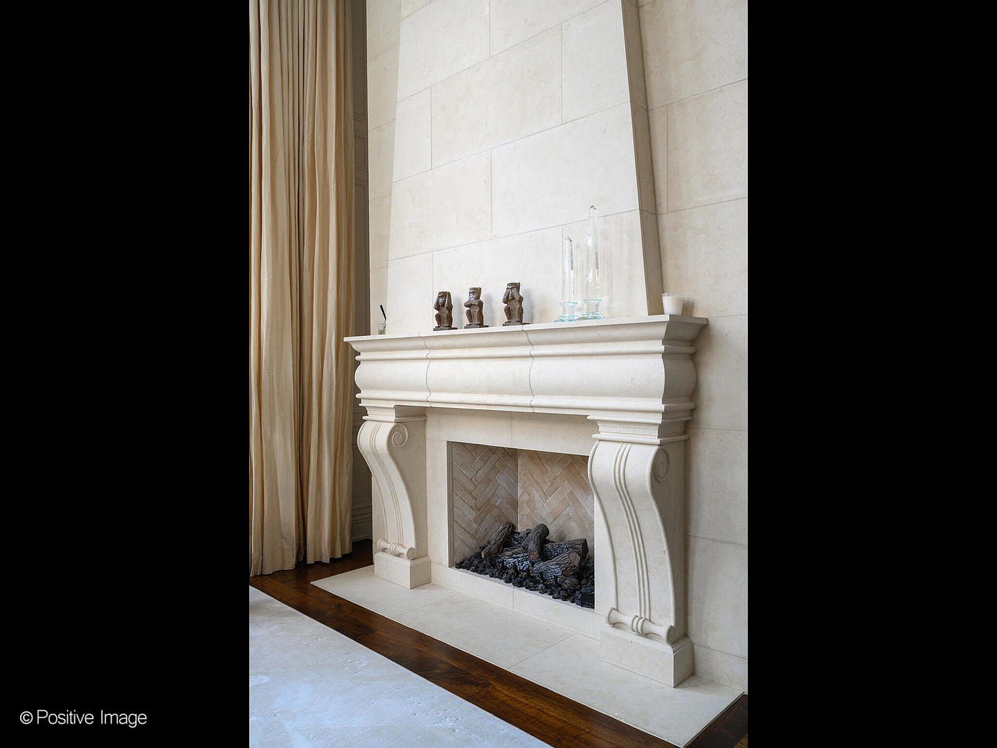 This image showcases a grand, custom-carved limestone fireplace mantel that serves as a sophisticated focal point in a high-end interior space. The fireplace features a classic herringbone-patterned brick firebox and is flanked by elegant, floor-to-ceiling beige curtains, emphasizing the room's vertical scale and luxurious aesthetic. The perspective is a straight-on, eye-level shot that highlights the intricate craftsmanship of the stone and the refined, neutral color palette of the room.