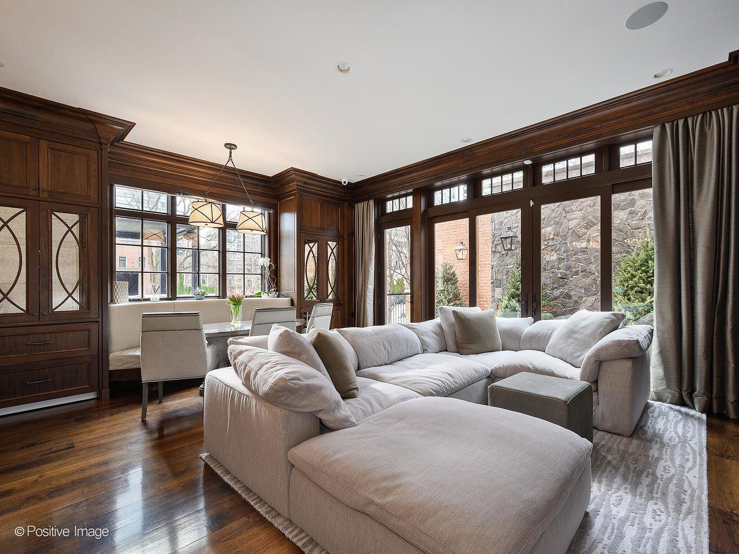 This spacious, open-concept living area features rich, dark wood paneling and flooring that creates a warm, sophisticated atmosphere. A large, plush sectional sofa serves as the centerpiece, complemented by a dining nook with built-in bench seating near the windows. The room is bathed in natural light from expansive glass doors and windows, offering a seamless transition to the outdoor stone patio.