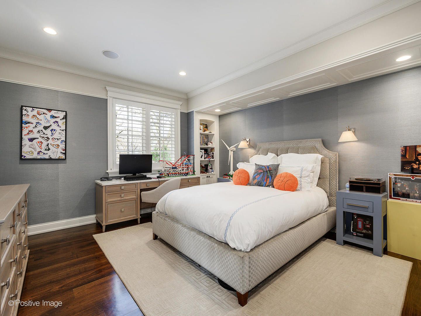 This well-appointed bedroom features a sophisticated gray color palette with textured wallpaper and dark hardwood flooring. The room is anchored by a plush upholstered bed with basketball-themed pillows, complemented by a built-in desk area and integrated shelving. The space is bright and inviting, showcasing a clean, modern aesthetic suitable for a youth or guest bedroom.