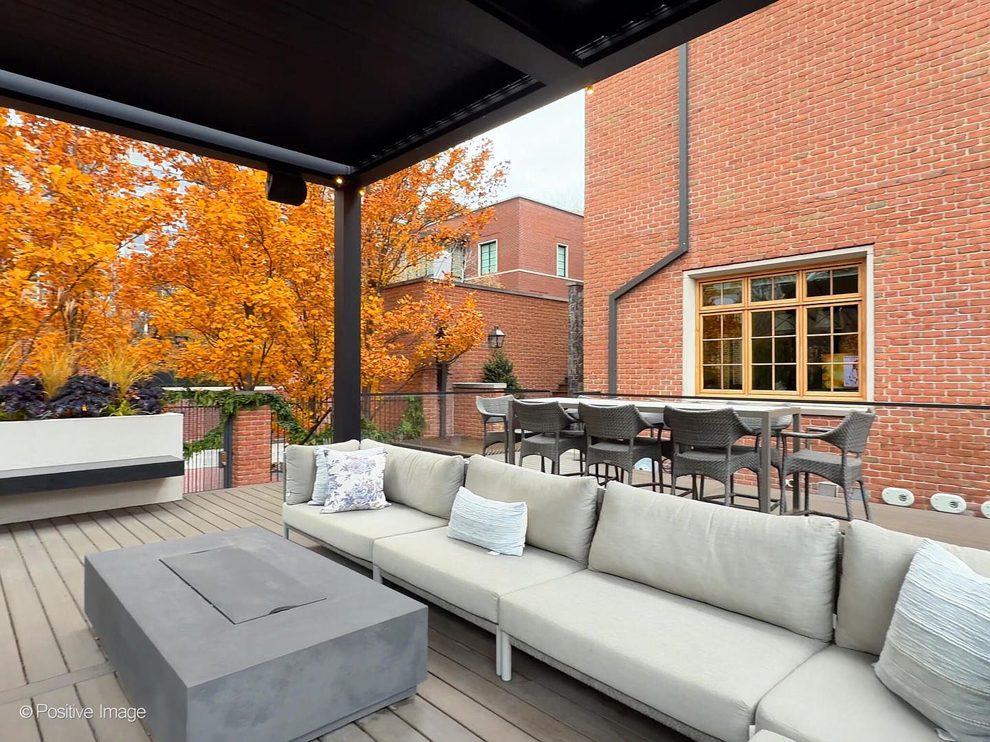 This sophisticated outdoor living space features a spacious deck equipped with a modern sectional sofa and a central concrete fire pit. A covered pergola provides shade, while a dining set sits nearby, all set against a backdrop of vibrant autumn foliage and a classic brick exterior. The perspective offers a welcoming, high-end atmosphere perfect for entertaining or relaxing in a private residential setting.