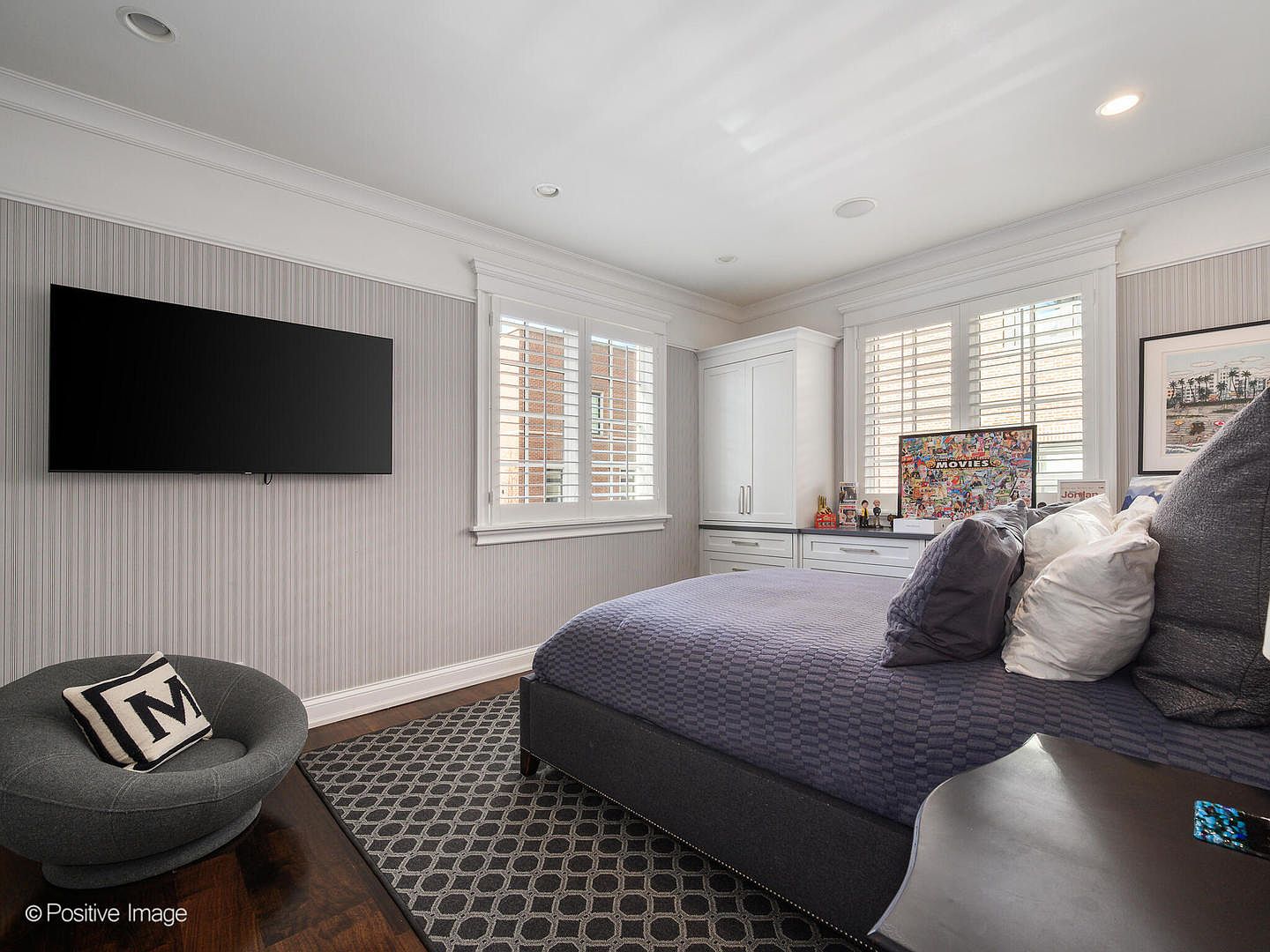 This cozy bedroom features a modern aesthetic with a dark blue patterned bedspread, a grey circular accent chair, and a large wall-mounted television. The room is accented by vertical striped wallpaper, white plantation shutters, and built-in cabinetry, creating a clean and organized living space. The perspective is from the corner of the room, capturing the bed, seating area, and storage solutions in a single, inviting frame.