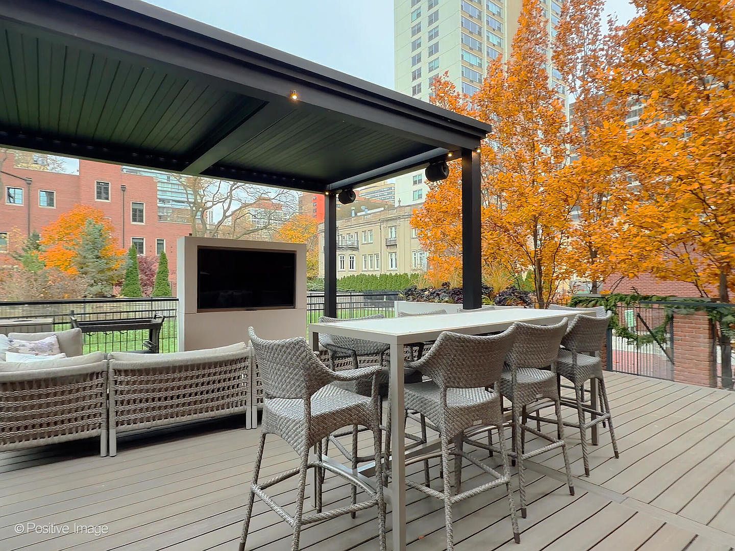 This sophisticated outdoor terrace features a covered dining and lounge area, complete with a sleek, modern pergola and a wall-mounted television. The space is furnished with high-quality wicker seating and a long dining table, set against a backdrop of vibrant autumn trees and urban architecture. The elevated perspective highlights a luxurious, well-appointed space perfect for entertaining in a city setting.