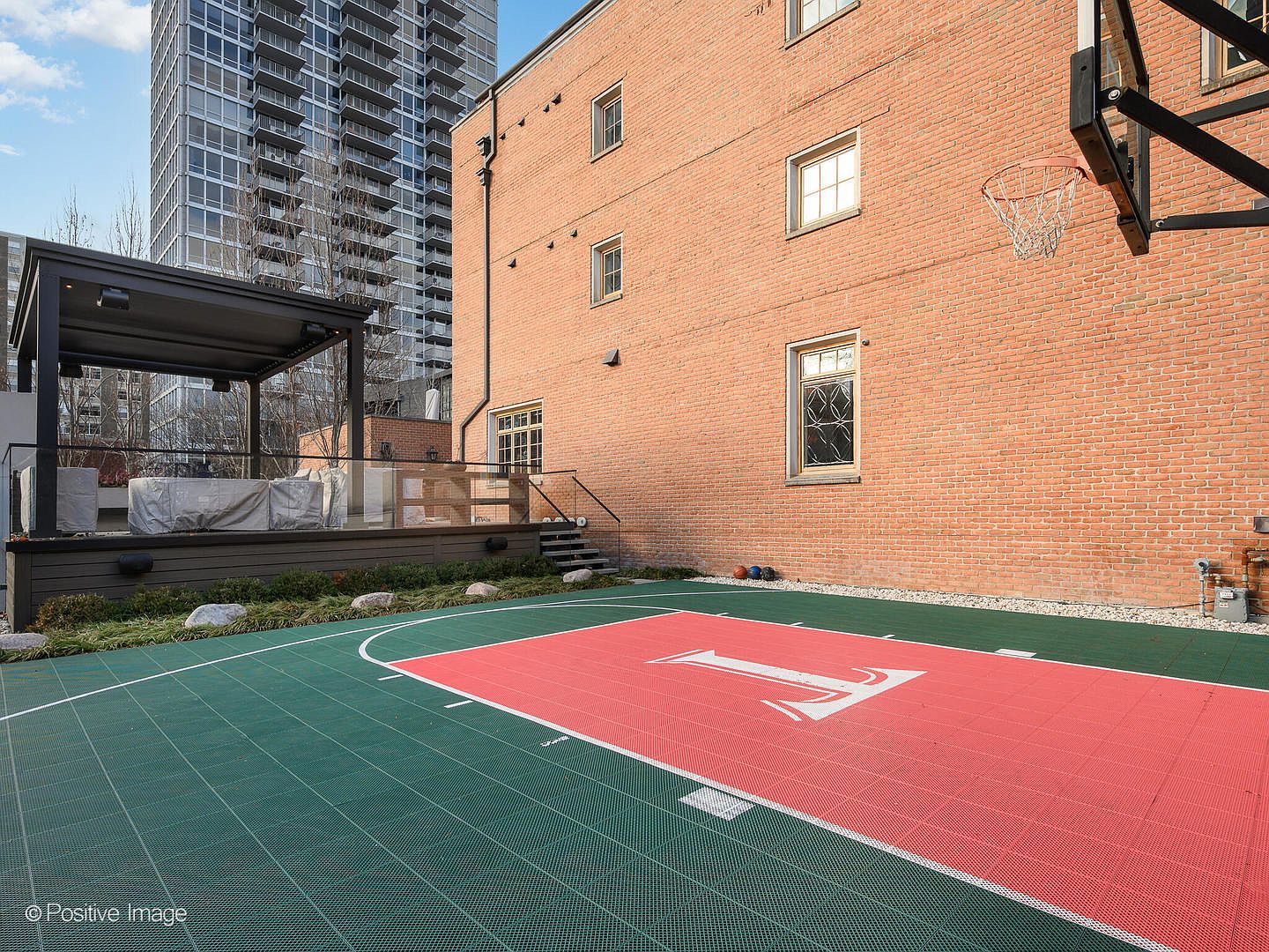 This outdoor space features a private basketball court with a vibrant red and green modular surface, complete with a basketball hoop mounted on the brick wall of the building. Adjacent to the court is a raised wooden deck with a modern pergola, providing a sophisticated area for outdoor lounging or entertaining. The scene is framed by a tall, contemporary high-rise building in the background, creating a striking contrast between the historic brick architecture and modern urban living.