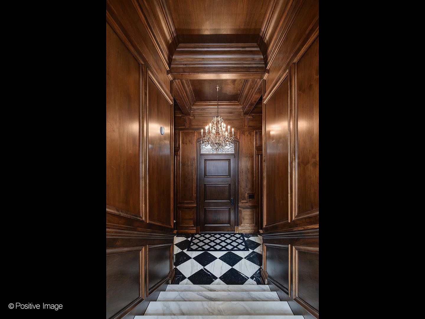 This grand entryway features rich, dark wood-paneled walls that extend from the floor to the ceiling, creating a sophisticated and traditional atmosphere. A striking black-and-white checkered marble floor leads to a heavy, dark wood door topped with an elegant transom window, all illuminated by a sparkling crystal chandelier. The perspective is captured from the top of a marble staircase, looking down into the foyer, emphasizing the architectural depth and luxurious craftsmanship of the home.