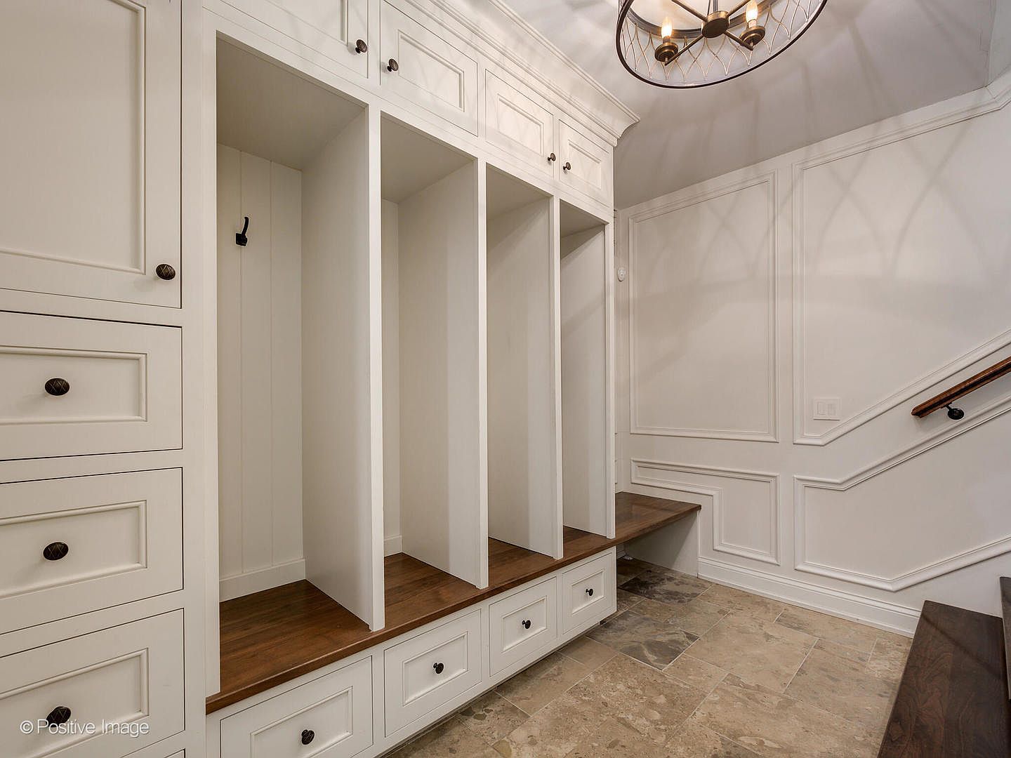 This elegant mudroom area features custom white built-in cabinetry with open cubbies, drawers, and a rich wood bench seat. The space is accented by sophisticated wall molding, stone tile flooring, and a stylish modern chandelier, creating a warm and organized entryway. The perspective captures the functional storage design alongside the transition to a staircase, highlighting a blend of luxury and practicality.