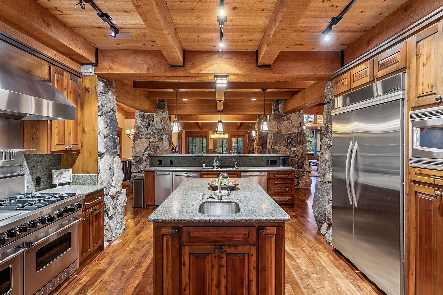 This is a spacious kitchen featuring wooden cabinetry, granite countertops, and stainless steel appliances. A large island with a sink is centrally located, and stone pillars add a rustic touch. The kitchen has a warm and inviting atmosphere with wooden beams on the ceiling.