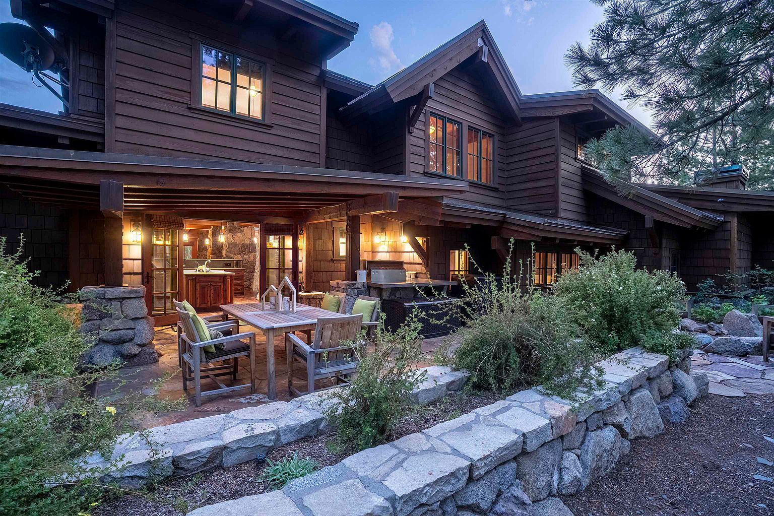 This image showcases a charming outdoor patio area attached to a rustic-style home. The patio features a wooden dining set with cushioned chairs, complemented by a stone-built outdoor kitchen area. The space is surrounded by lush greenery and a stone retaining wall, creating a cozy and inviting atmosphere perfect for outdoor entertaining.