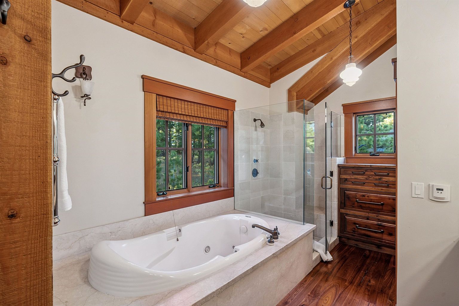 This is a primary bathroom featuring a large soaking tub with marble surround, a glass-enclosed shower, and wood-framed windows with woven shades. The room has a rustic-chic style with exposed wooden beams on the vaulted ceiling and dark wood flooring. A wooden dresser provides storage, and the walls are painted in a neutral tone.