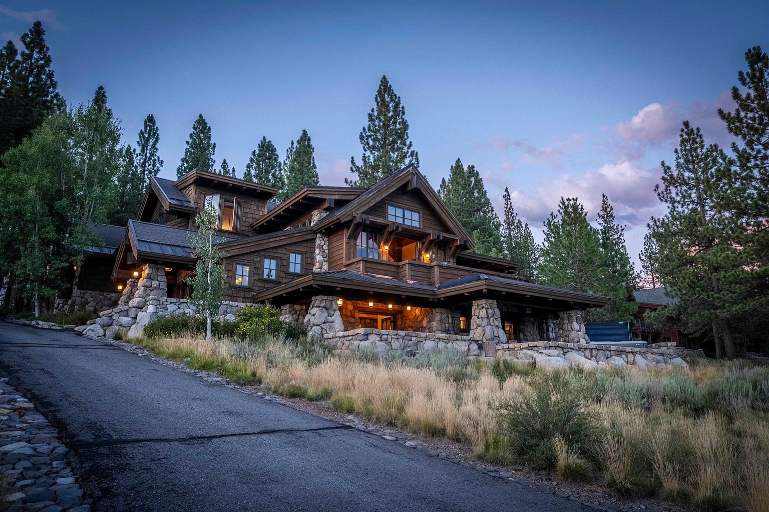 This is a captivating front view of a luxurious mountain home, showcasing its impressive stone foundation and timber frame construction. The multi-level design features a covered porch with stone pillars, creating an inviting entryway. The surrounding landscape includes mature trees and natural grasses, enhancing the property's serene and private setting.