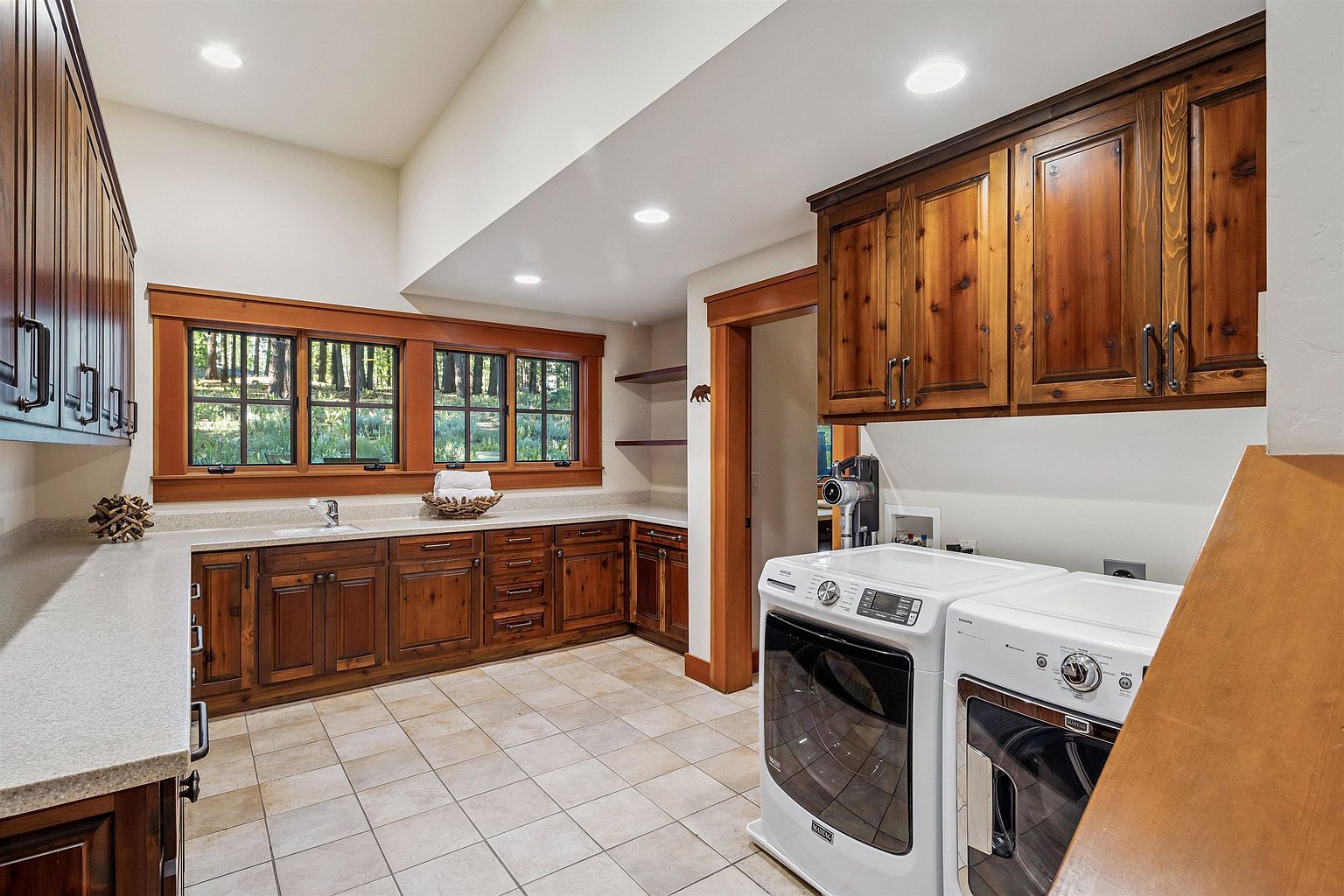This is an interior shot of a laundry room featuring wooden cabinetry and trim, a light-colored countertop, and a pair of front-loading washing machines. The room has a warm and inviting feel, with natural light coming in through the windows above the sink. The tile flooring and recessed lighting add to the room's functionality and aesthetic appeal.