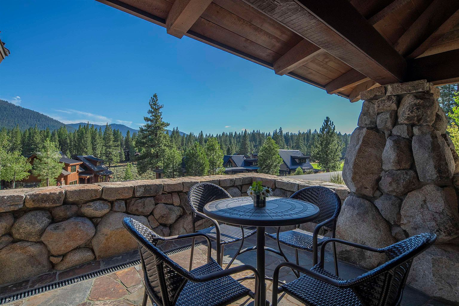 This image showcases a charming outdoor patio or balcony area, featuring a stone wall and pillar construction that adds a rustic touch. A round table with four wicker chairs is centered on the patio, suggesting a cozy space for relaxation or dining. The background reveals a scenic view of trees and houses, enhancing the property's appeal.