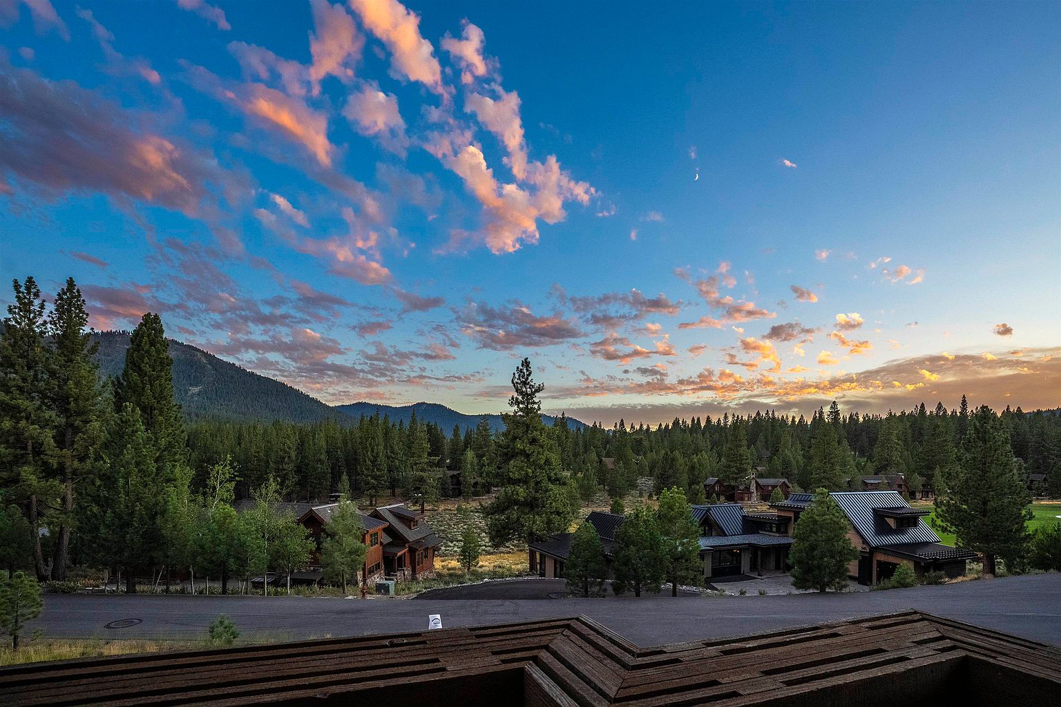 This image showcases a picturesque front view of several residential properties nestled within a wooded landscape. The homes feature a blend of modern and rustic architectural styles, with dark roofs and wood accents. The scene is set against a backdrop of lush trees, mountains, and a vibrant sky with scattered clouds, creating a serene and inviting atmosphere.