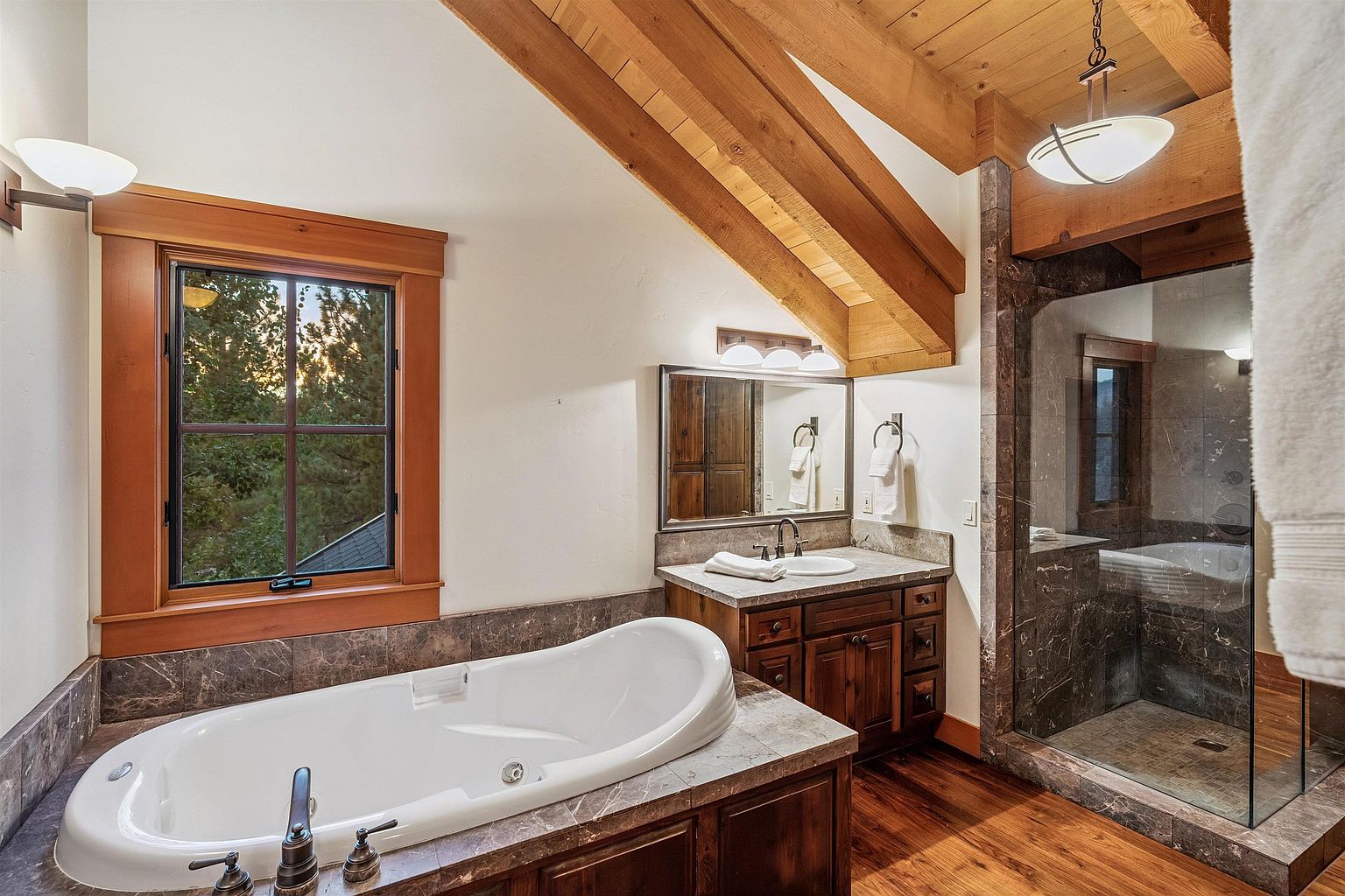 This is a primary bathroom featuring a soaking tub with dark marble surround, a wooden vanity with a marble countertop, and a glass-enclosed shower. The bathroom has a rustic-modern style with exposed wooden beams on the ceiling and hardwood floors. The space is well-lit with natural light from a window and artificial lighting from fixtures above the mirror and shower.