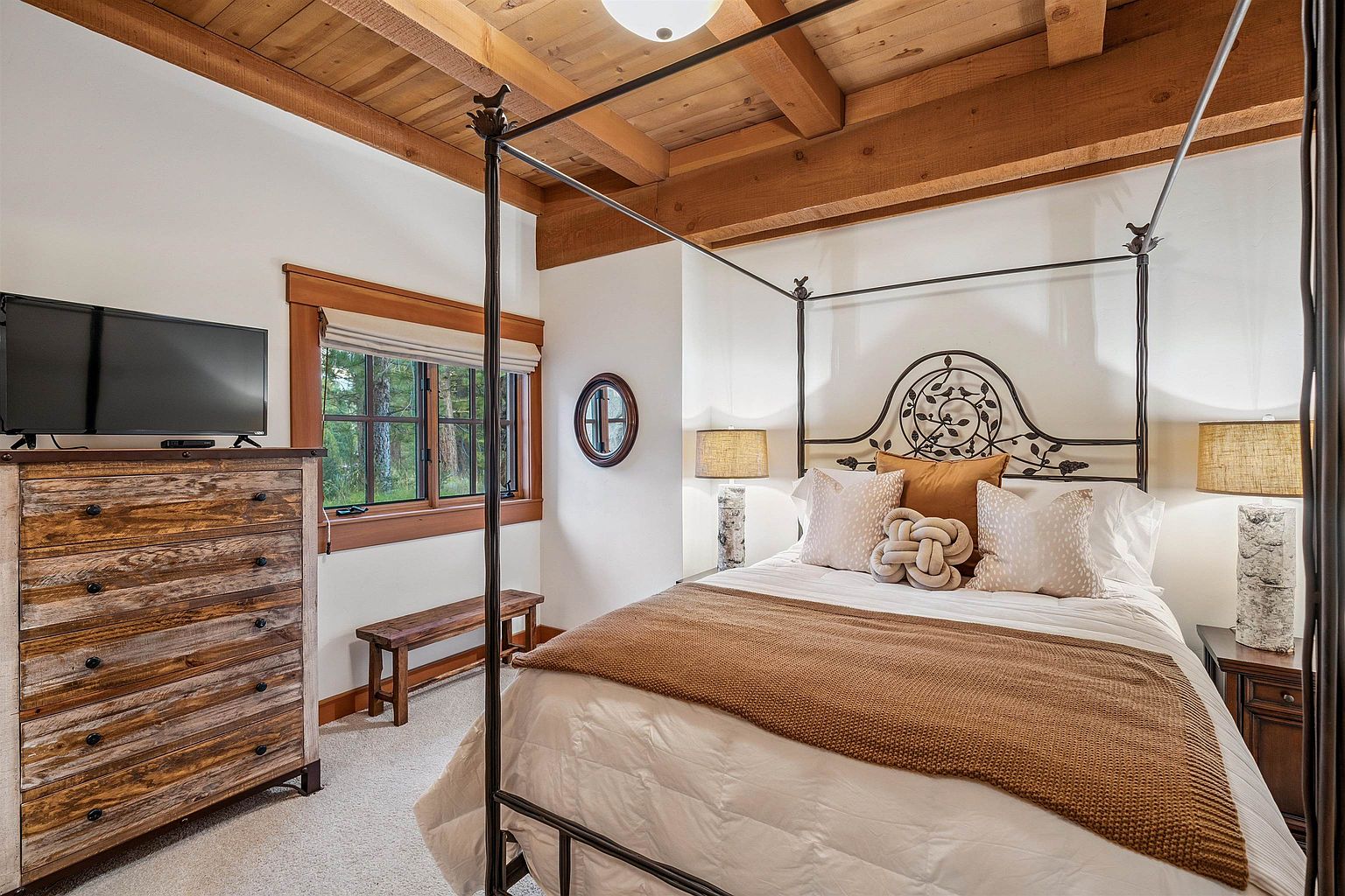This is a cozy primary bedroom featuring a four-poster bed with decorative ironwork, complemented by warm-toned bedding and pillows. A rustic wooden dresser with a television sits against one wall, while a window provides natural light and a view of the outdoors. The room has a warm and inviting atmosphere with a blend of rustic and elegant elements.