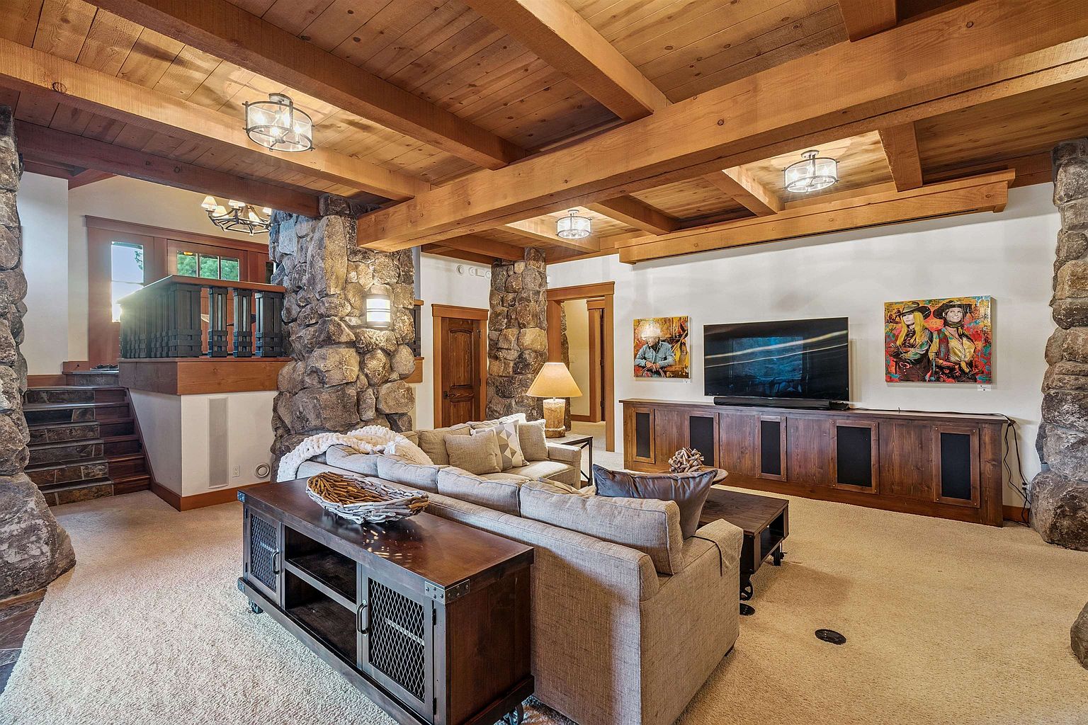 This is an interior shot of a living room featuring a stone pillar, wooden beam ceiling, and a large sectional sofa. A wooden entertainment center houses a flat-screen TV, and artwork adorns the walls. The room exudes a rustic yet comfortable atmosphere, perfect for relaxation and entertainment.
