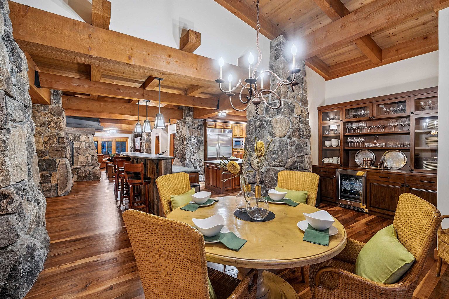 This is an interior shot of a dining room featuring a round wooden table set for four, surrounded by wicker chairs with green cushions. A stone pillar and wooden beams add rustic charm, while a built-in cabinet displays glassware and a wine cooler. The room exudes a warm and inviting atmosphere, perfect for family gatherings.