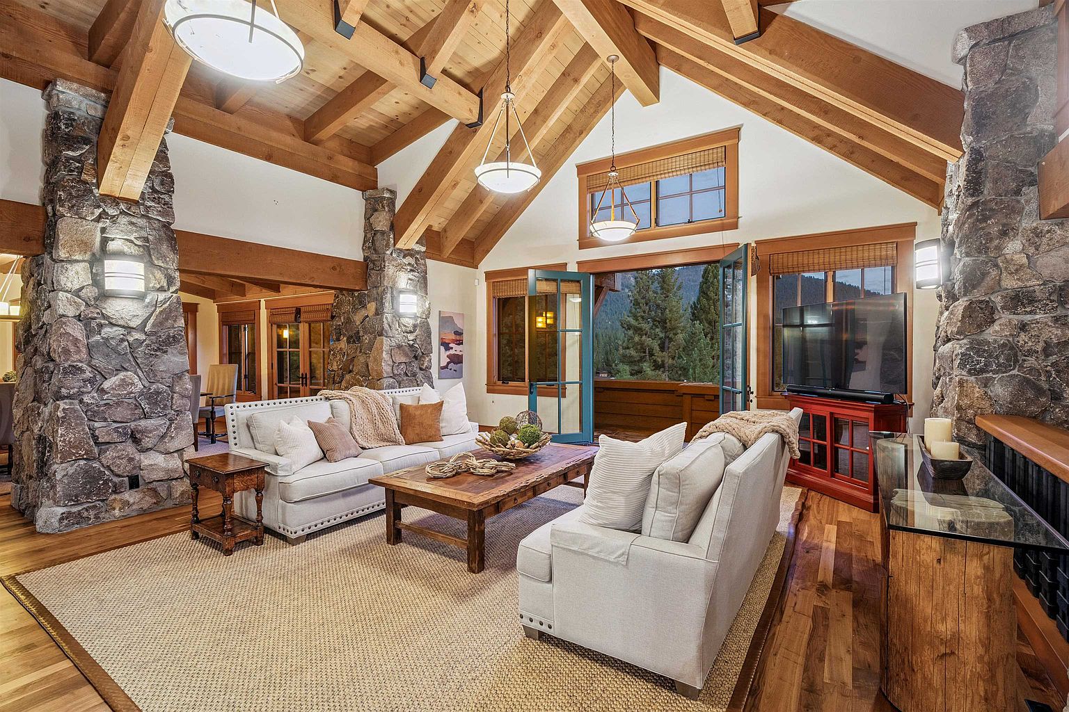 This is a warm and inviting living room featuring a vaulted ceiling with exposed wooden beams and stone pillars. The room is furnished with comfortable sofas, a rustic wooden coffee table, and a large area rug. Natural light floods the space through large windows and doors, offering views of the surrounding landscape.