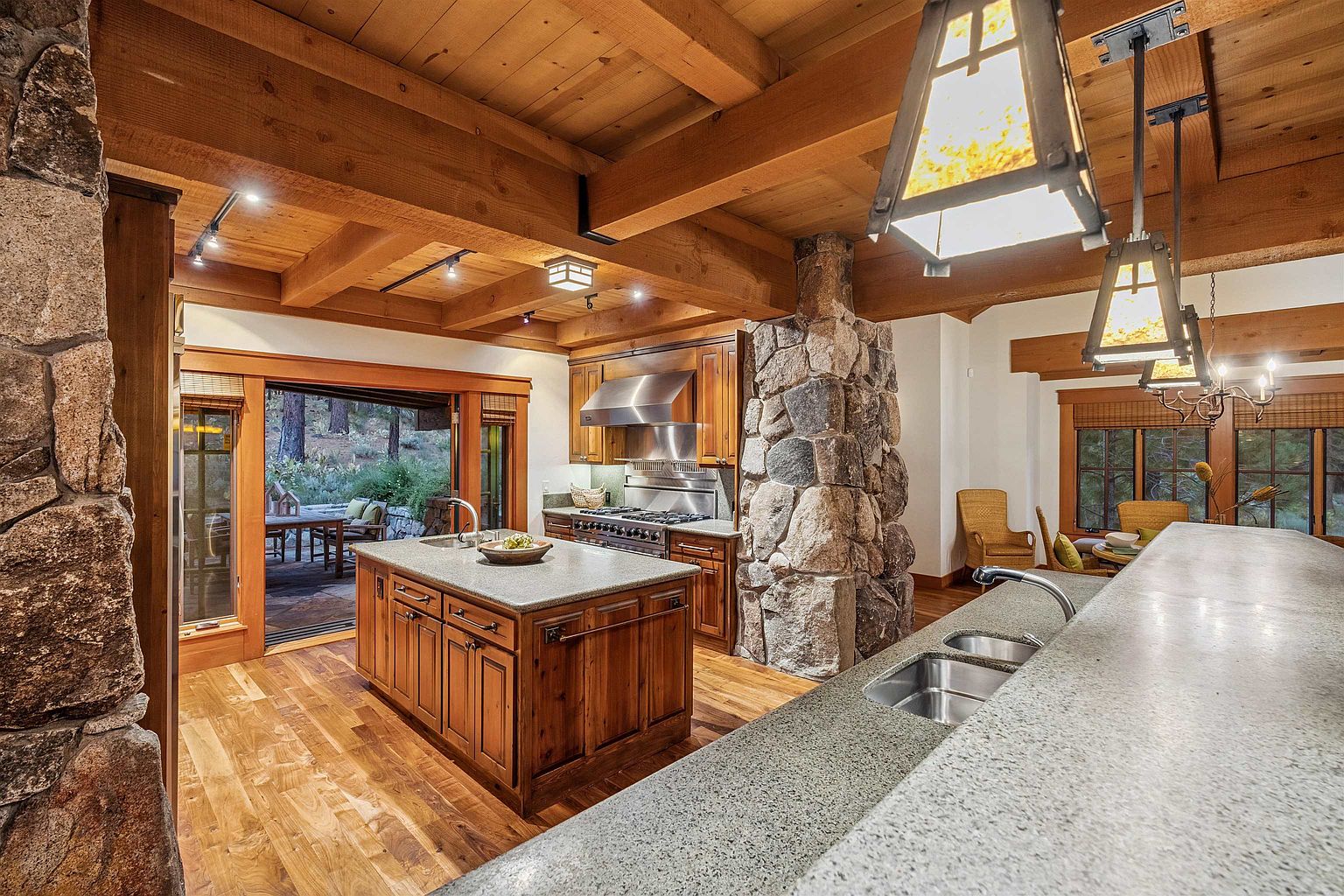This is a warm and inviting kitchen featuring wood cabinetry, granite countertops, and a stone accent wall. The kitchen island provides ample workspace, and the open layout connects to an outdoor patio area. The exposed wood beam ceiling and unique lighting fixtures add to the rustic charm of the space.