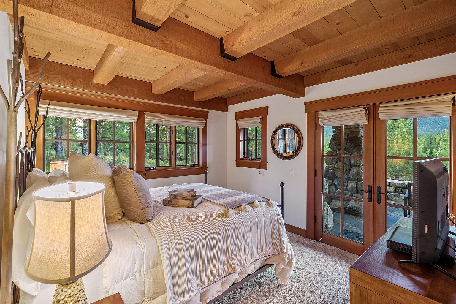 This is a cozy primary bedroom featuring a wooden beamed ceiling and multiple windows that offer views of the surrounding trees. The bed is neatly made with neutral-toned bedding, and a bedside lamp provides soft lighting. A round mirror hangs on the wall, and a set of French doors leads to an outdoor area, enhancing the room's connection to nature.