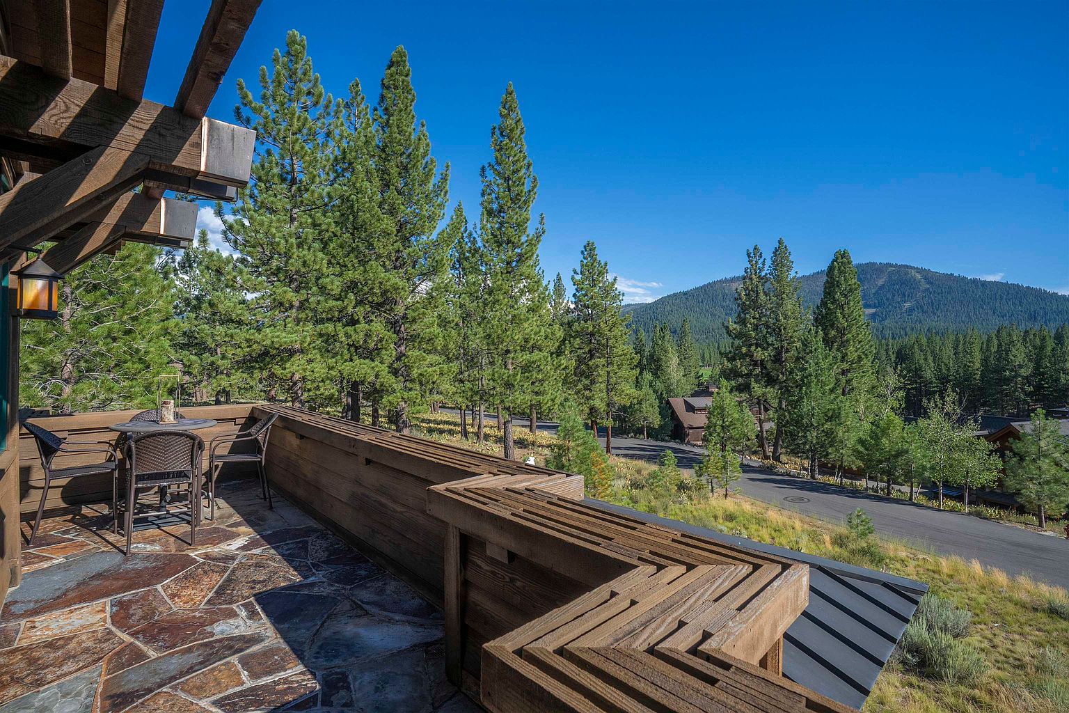 This image showcases a charming patio/deck area with a stone tile floor and a unique wooden railing design. A small table and chairs are set up, suggesting a cozy outdoor seating area. The view extends to a landscape of trees and a distant mountain, creating a serene and inviting atmosphere.