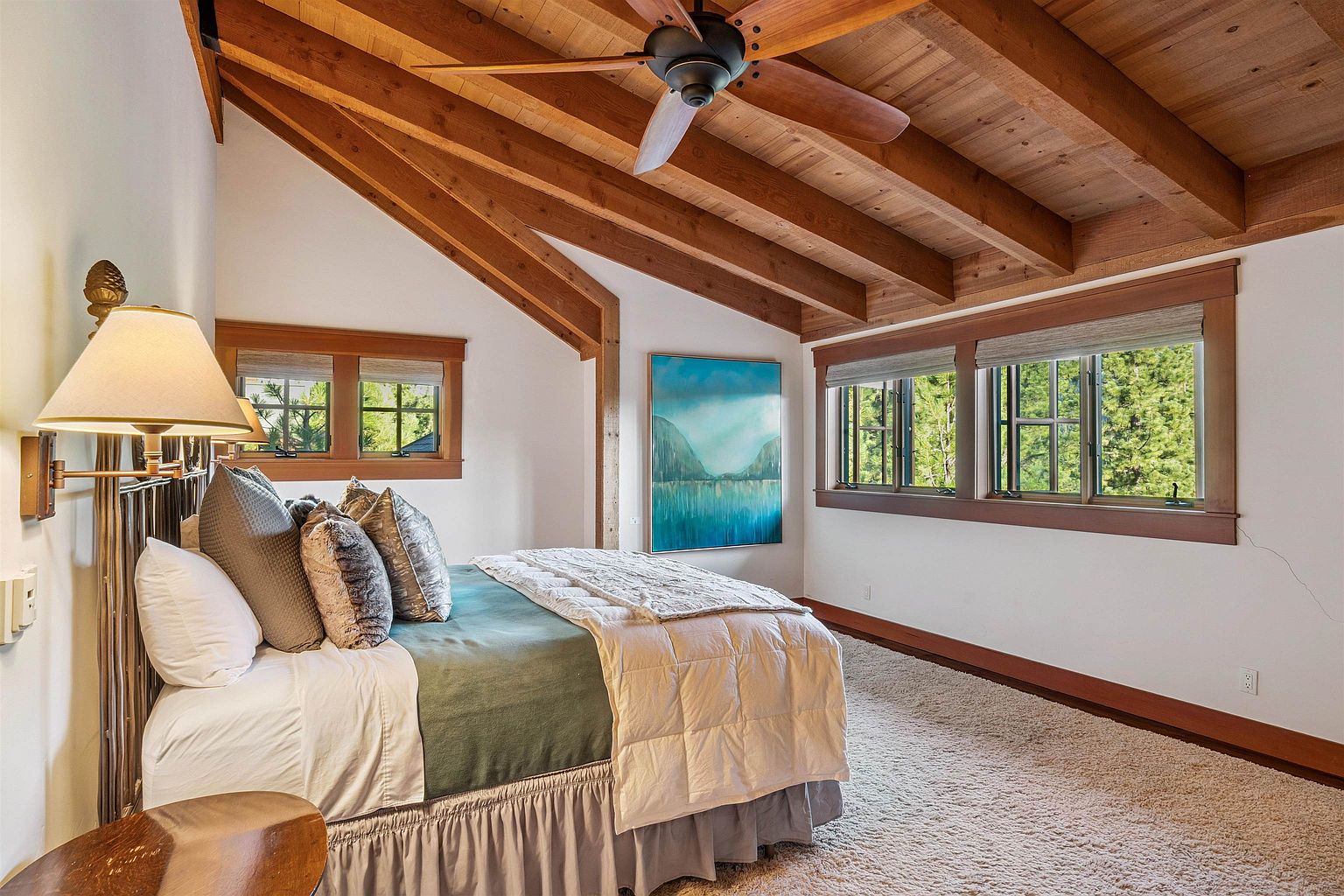 This primary bedroom features a vaulted wood beam ceiling and a large bed with multiple pillows and a patterned comforter. Natural light streams in through the windows, highlighting the neutral color palette and cozy atmosphere. A painting hangs on the wall, adding a touch of color to the room.