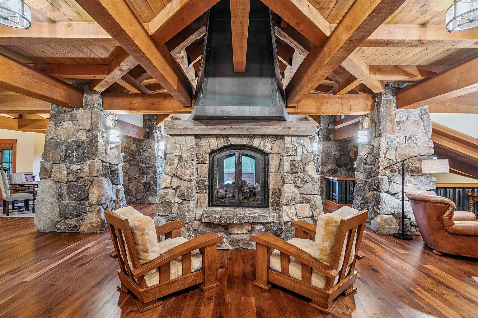 This interior shot showcases a rustic living room with a grand stone fireplace as its focal point. The room features exposed wooden beams, stone pillars, and hardwood floors, creating a warm and inviting atmosphere. Two wooden armchairs are positioned in front of the fireplace, suggesting a cozy seating area.