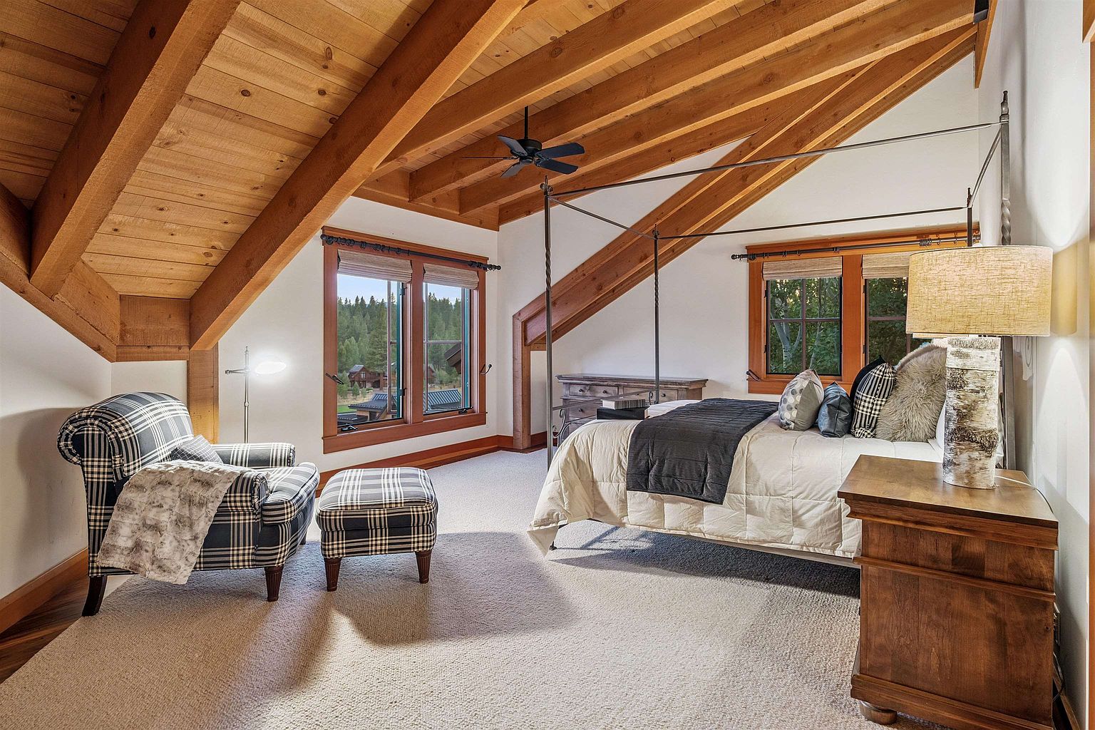 This is a cozy primary bedroom featuring a vaulted wood ceiling with exposed beams, creating a rustic and inviting atmosphere. The room is furnished with a four-poster bed, a plaid armchair and ottoman, and wooden nightstands, complemented by natural light streaming through the windows. The overall impression is one of warmth and comfort, perfect for a relaxing retreat.