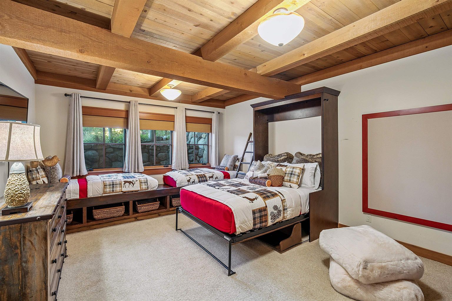 This is a cozy guest bedroom featuring a rustic design with wooden beams on the ceiling and built-in window seats. The room includes a Murphy bed and additional sleeping spaces, making it ideal for accommodating multiple guests. The decor incorporates natural elements and warm tones, creating a welcoming and comfortable atmosphere.