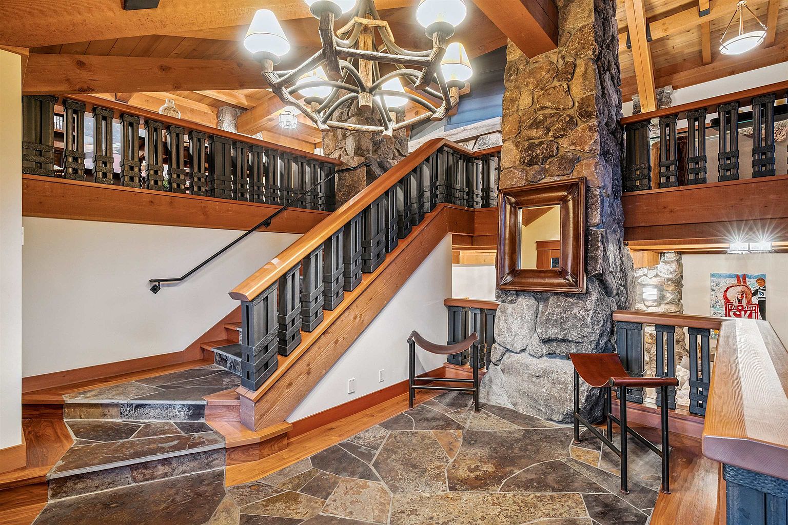This interior shot showcases a grand hallway and staircase featuring a blend of rustic and elegant design elements. The staircase has wooden steps and black metal railings, complemented by a stone accent wall and a unique chandelier. The flooring is a mix of wood and stone tiles, adding to the luxurious feel of the space.