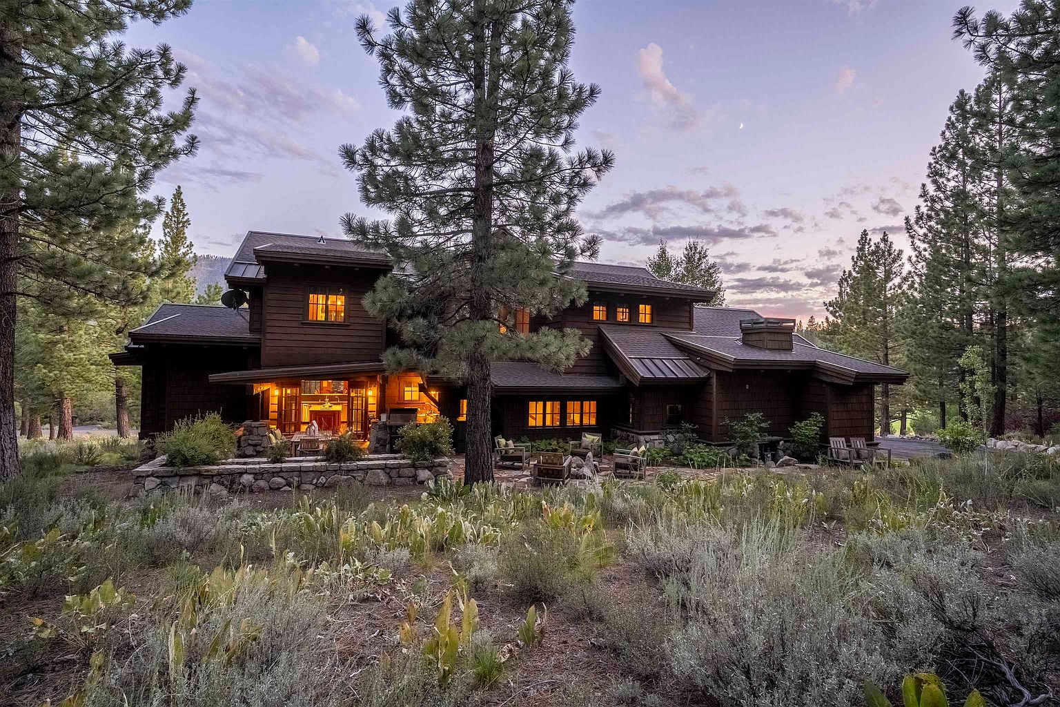 This image showcases the front exterior of a large, multi-story home with a rustic, lodge-like aesthetic. The house features dark wood siding, a stone foundation, and multiple windows that glow warmly, suggesting a cozy interior. A mature pine tree stands prominently in the foreground, adding to the natural, secluded feel of the property, while a stone patio with outdoor seating invites relaxation.