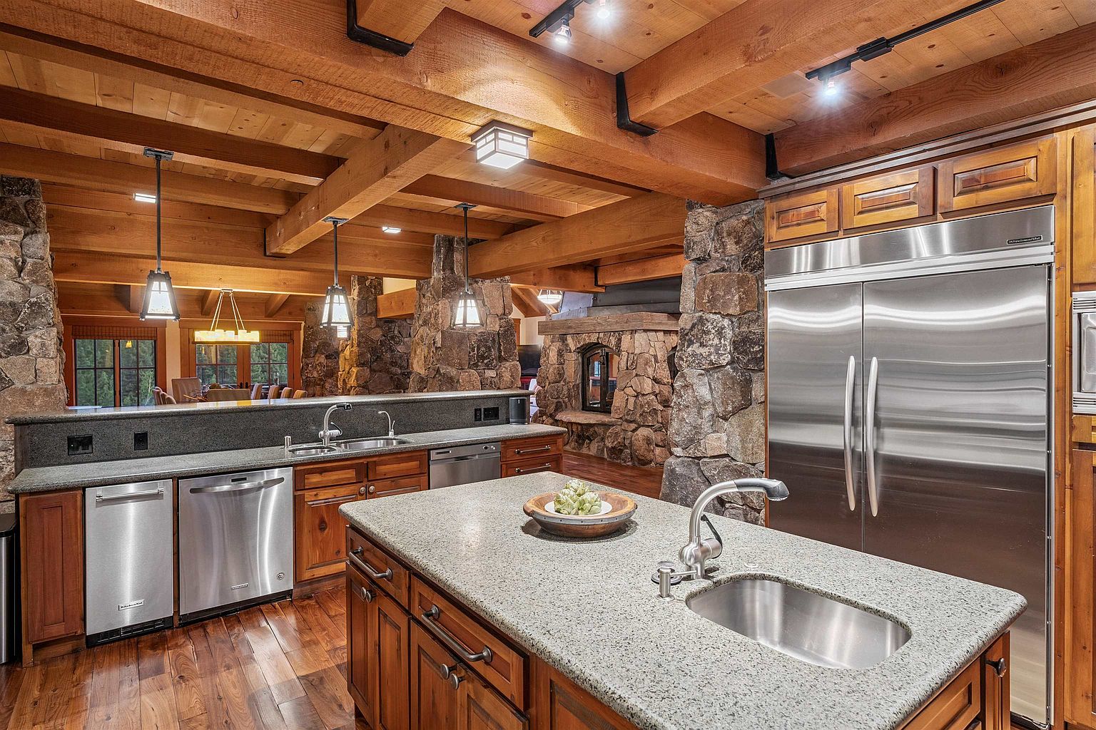 This is a warm and inviting kitchen featuring wooden cabinetry, granite countertops, and stainless steel appliances. The kitchen has a rustic charm with exposed wooden beams and stone accents. A large island with a sink provides ample workspace, and the overall impression is one of comfort and functionality.